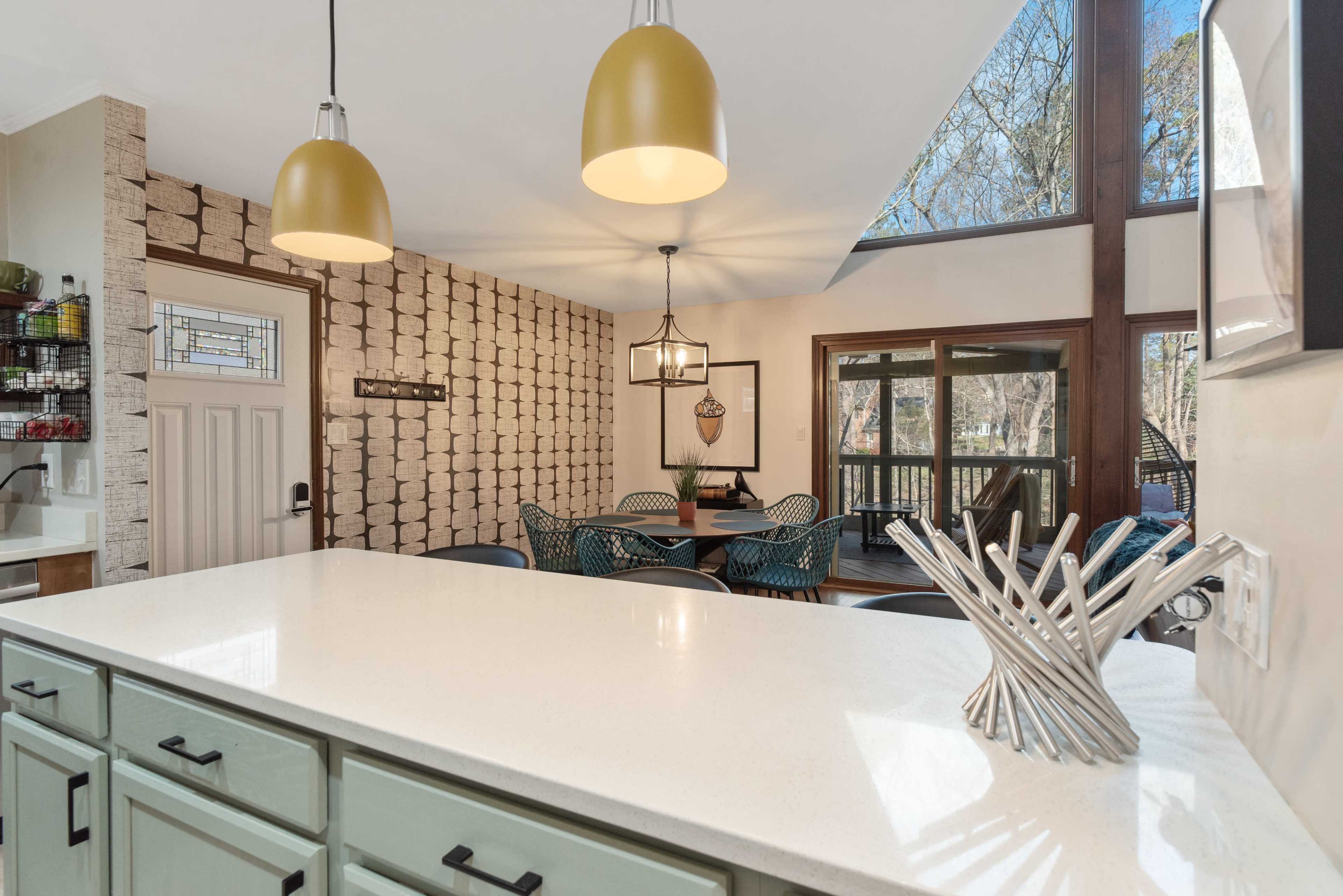 A modern kitchen features a large island with a white countertop, pendant lights, and a dining area with a view of outdoor greenery.