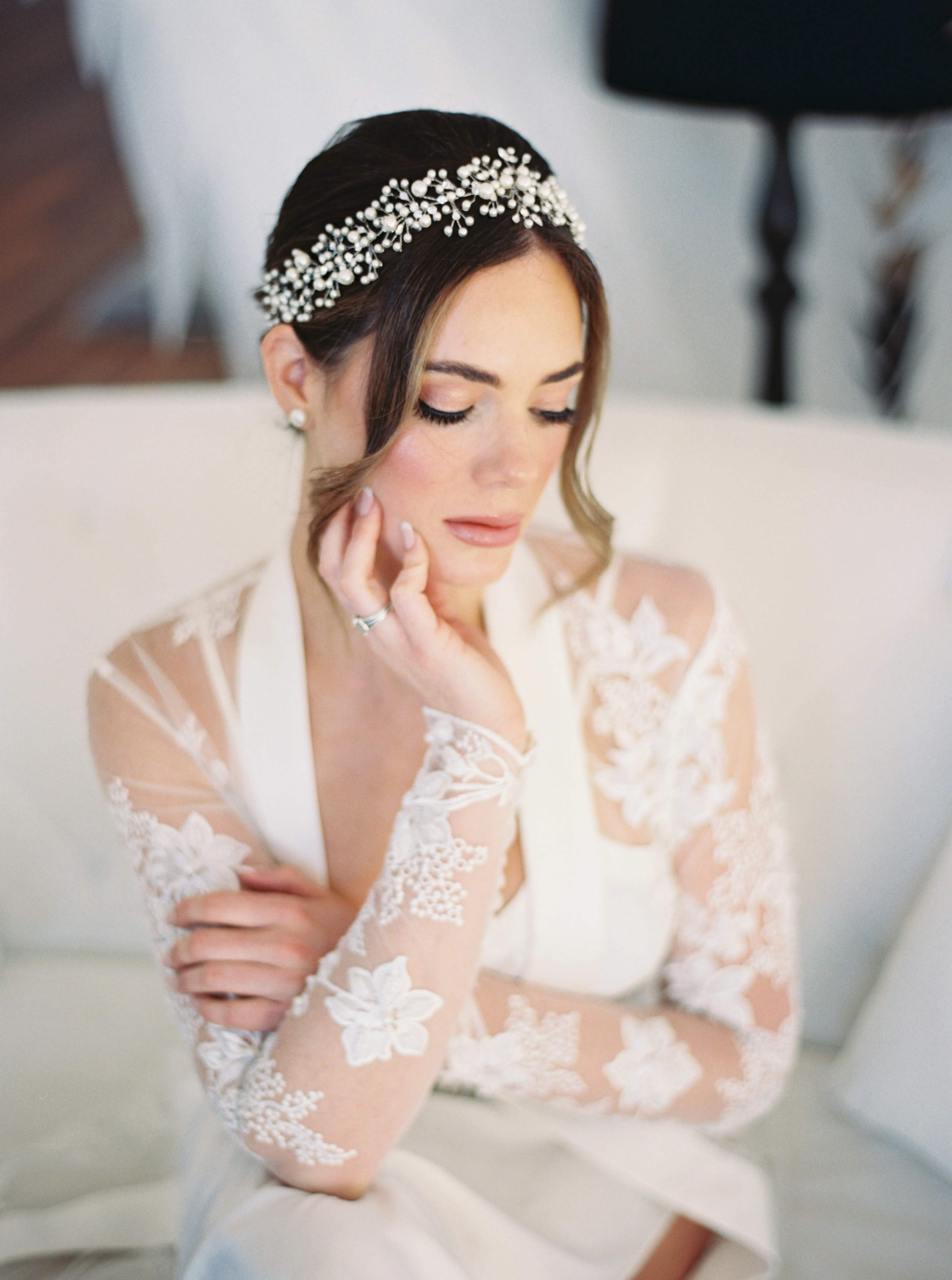A woman in a detailed lace wedding dress sits thoughtfully on a white sofa, wearing a jeweled hairpiece.