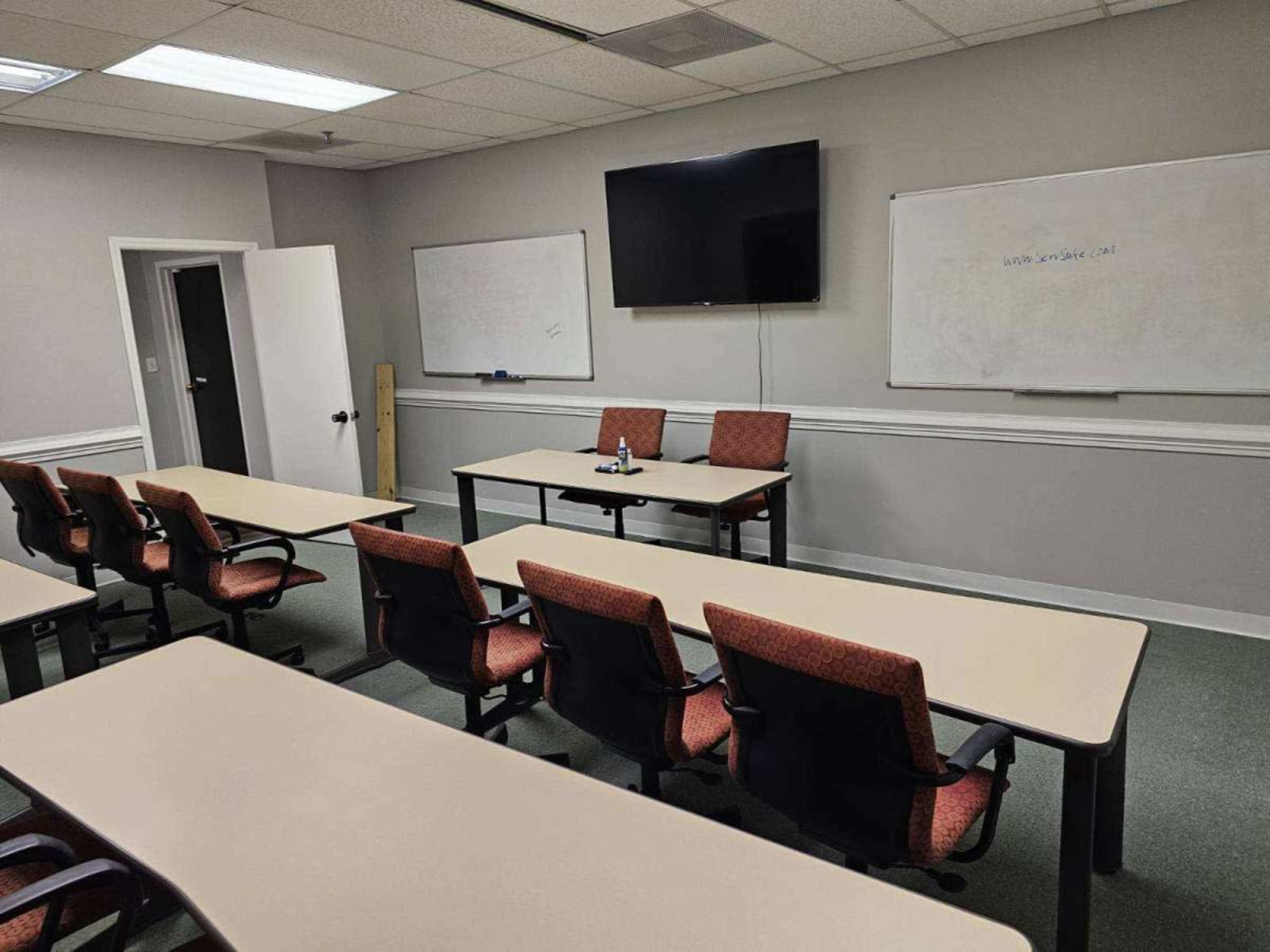 A conference room with several rows of tables and chairs, featuring a wall-mounted TV and whiteboards.