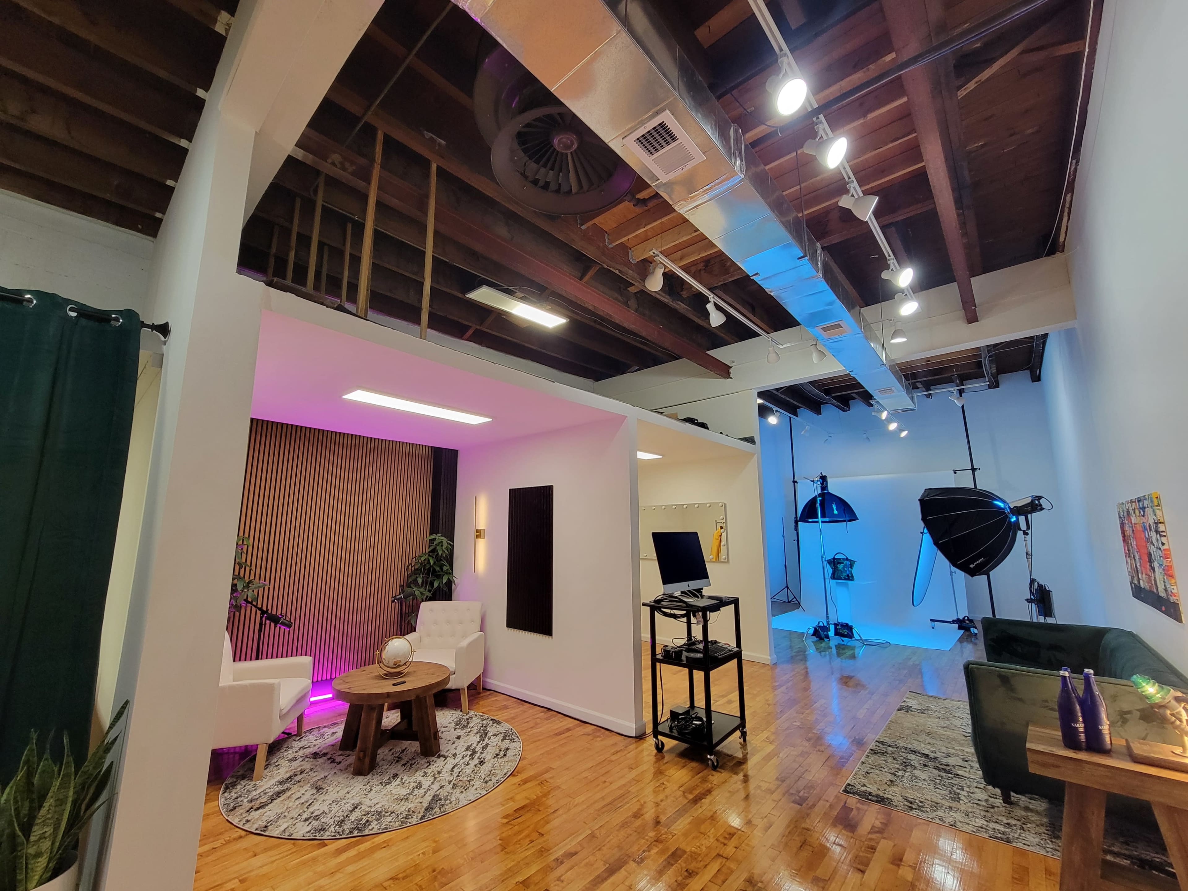 The image shows a modern studio space divided into two areas, one featuring a seating arrangement with white chairs and a wooden table, and the other equipped with professional lighting and photography equipment.