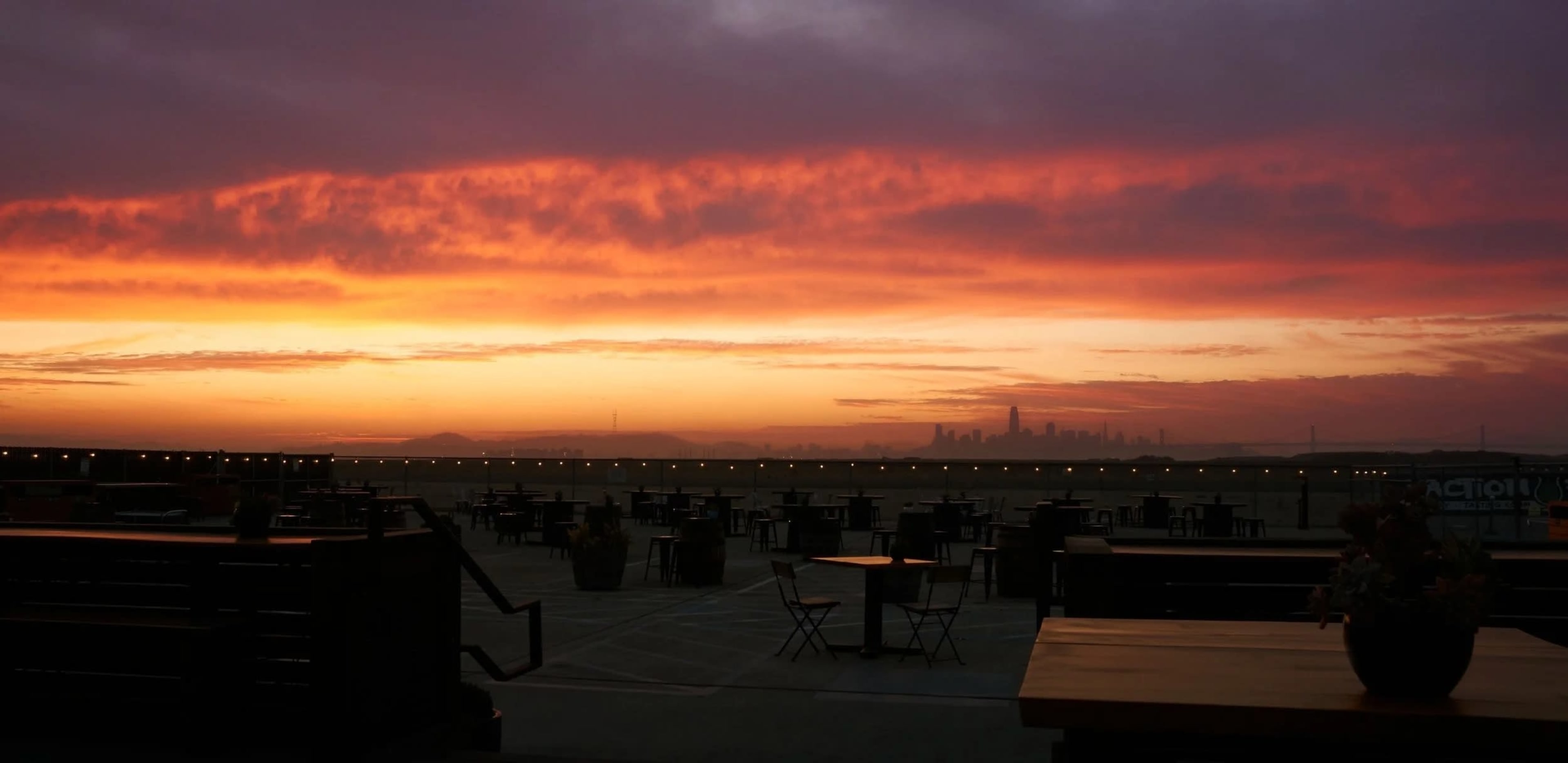 The image shows a sunset with vibrant colors behind a city skyline, framed by empty tables and chairs in the foreground.