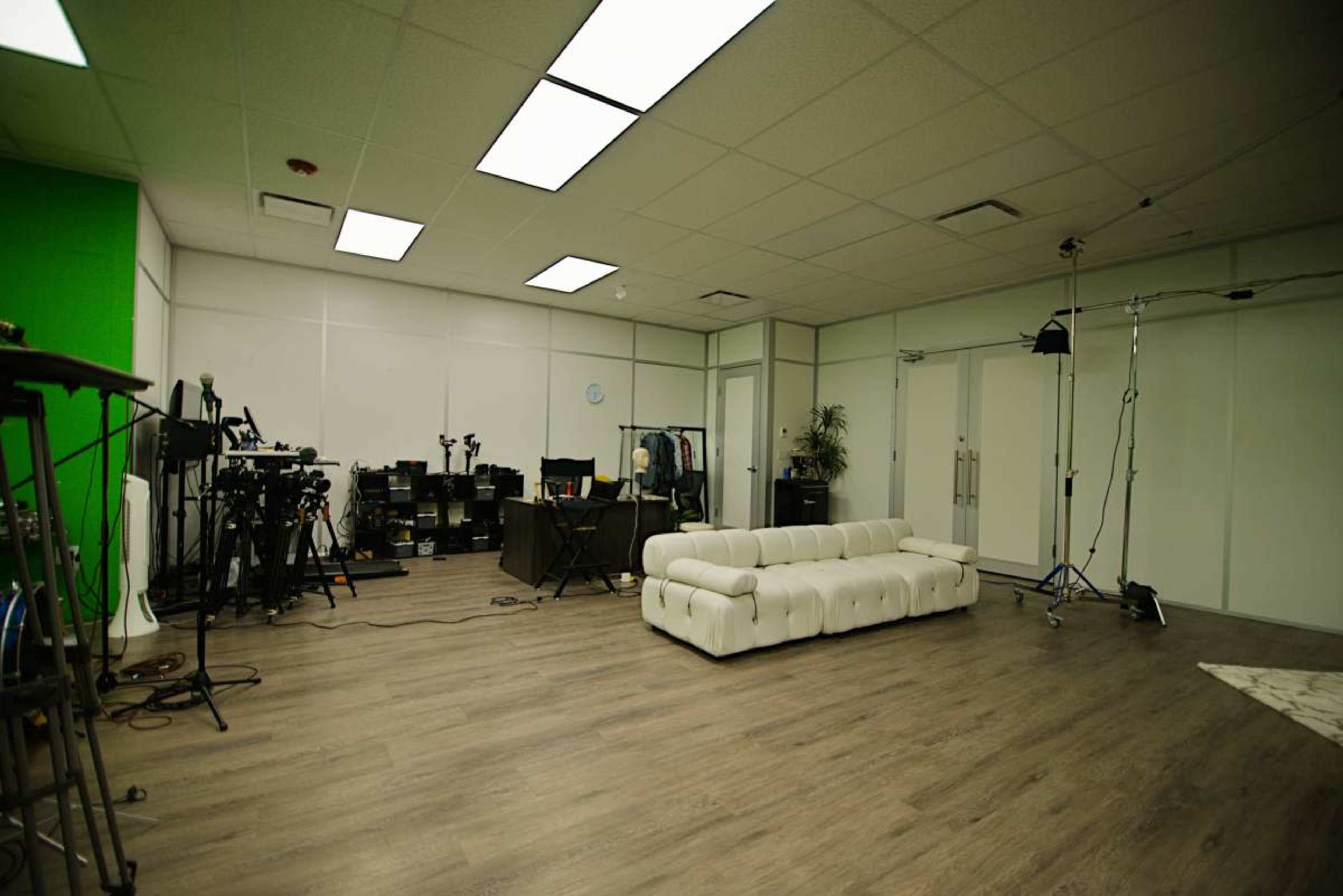 The image shows a sparse studio space featuring a white sofa, several cameras on tripods, equipment, and a green screen on one wall.