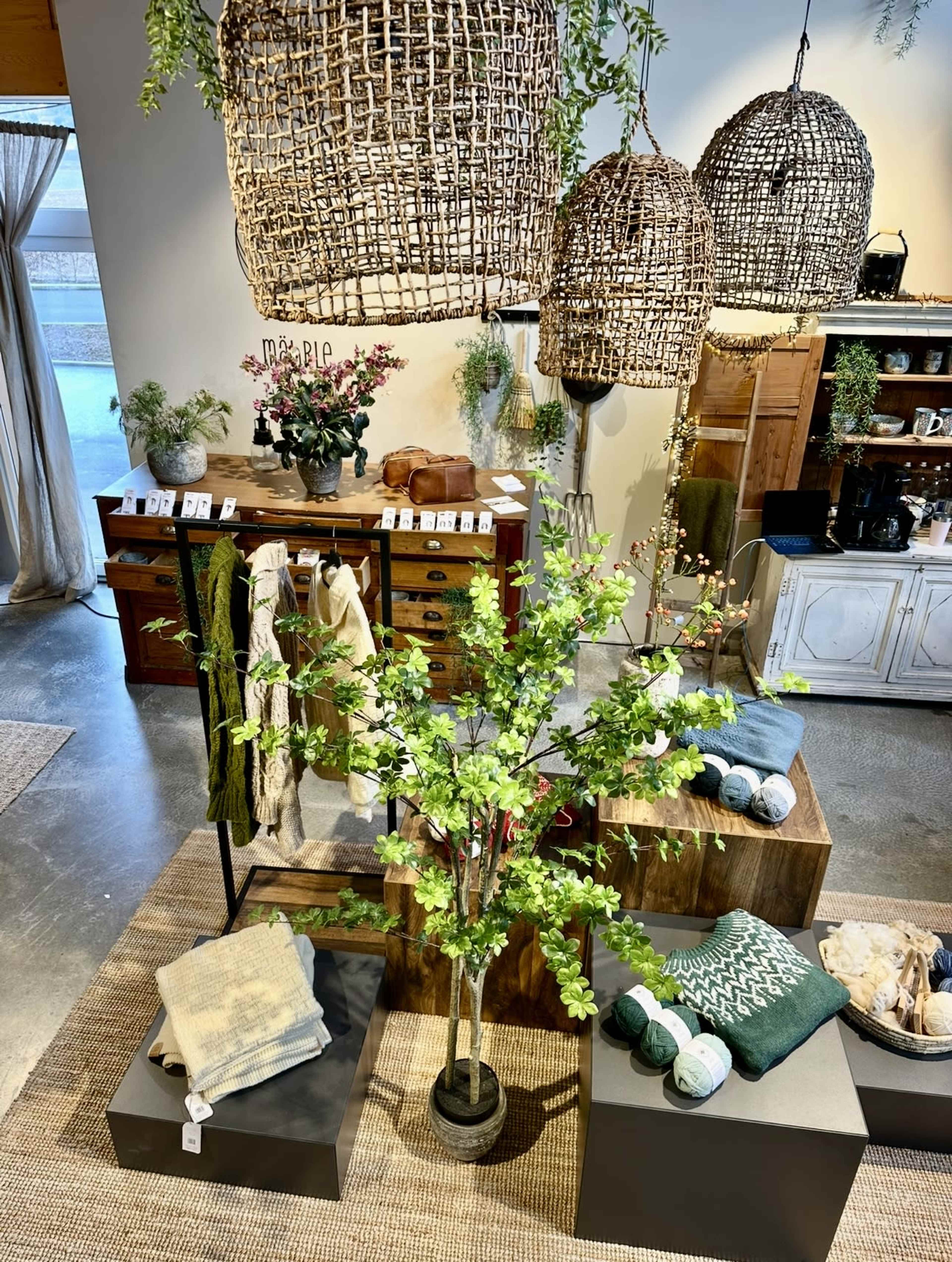 The image shows a home decor store display featuring woven pendant lights, wooden furniture, greenery, and various textiles arranged on tables.