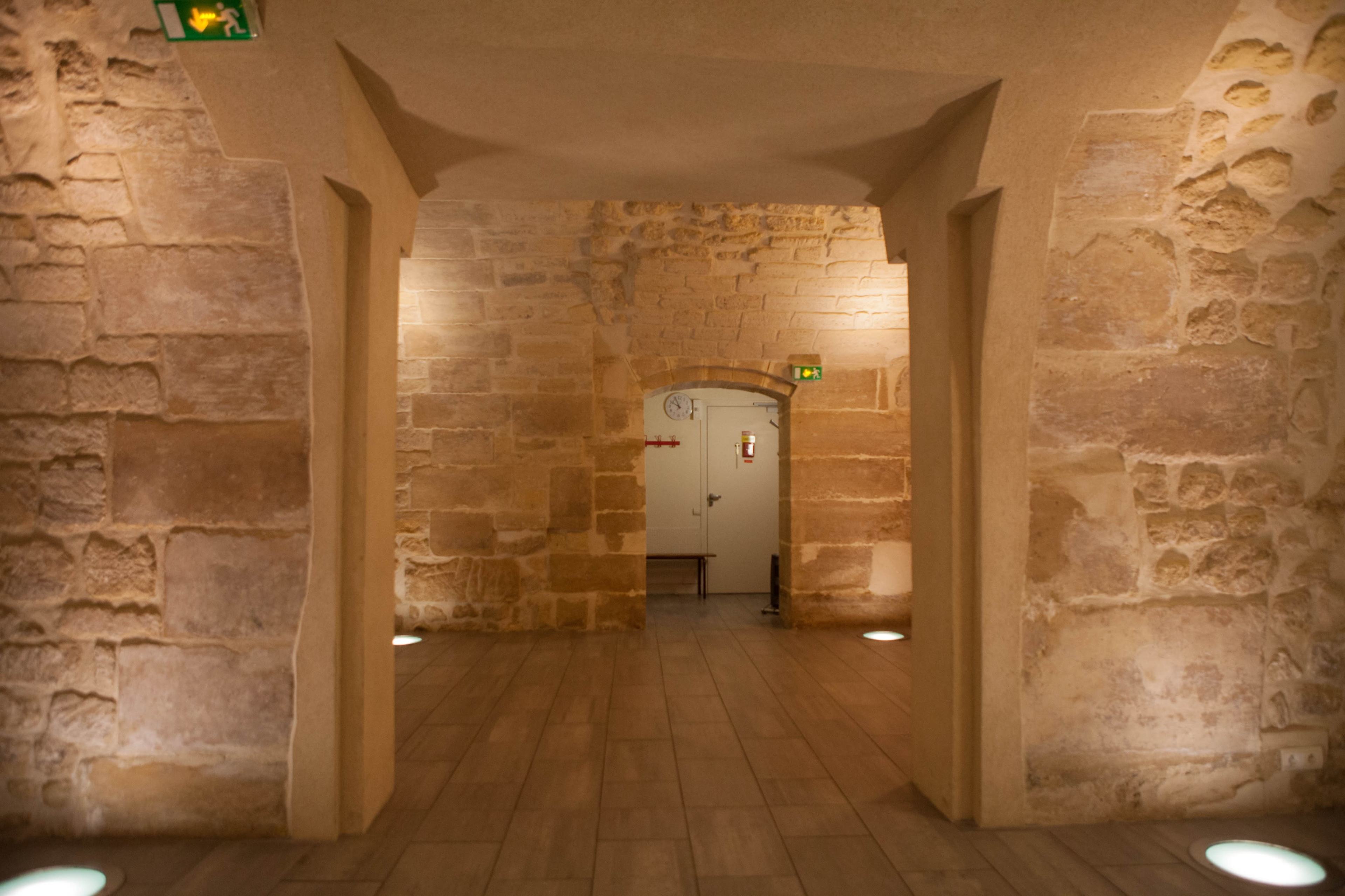 The image shows a corridor with stone walls and wooden flooring, leading to an arched opening and a door in the background.