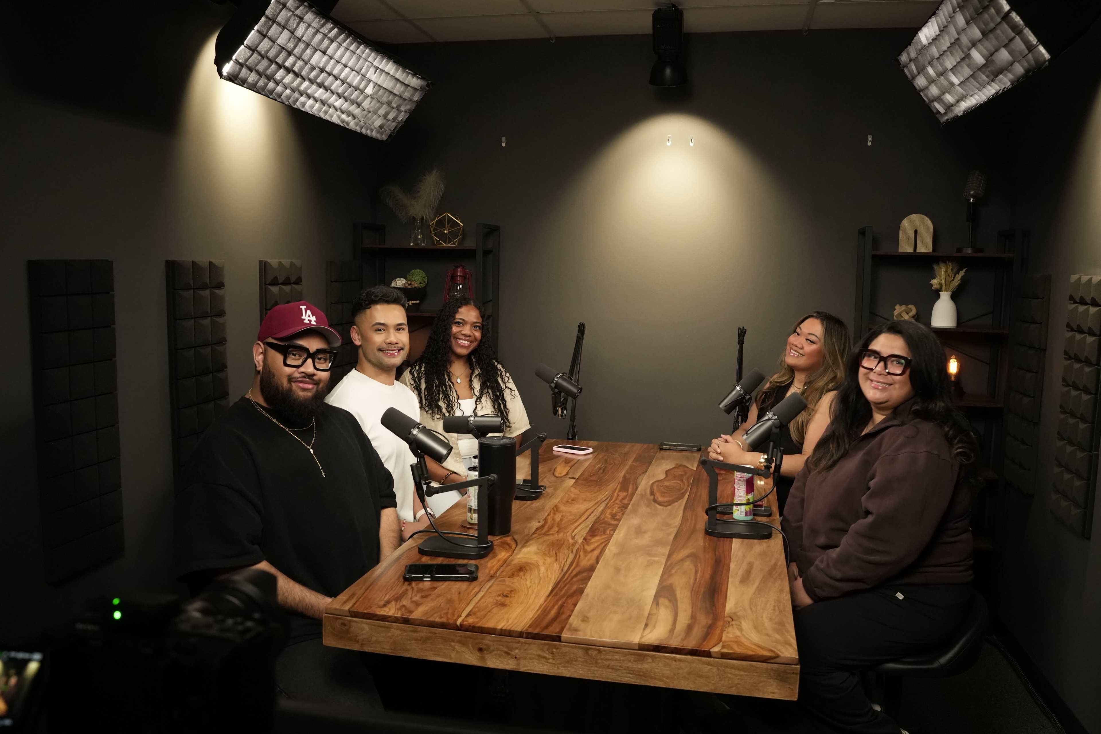 A group of five people is seated at a wooden table in a podcast studio, each with a microphone in front of them, ready for a discussion.