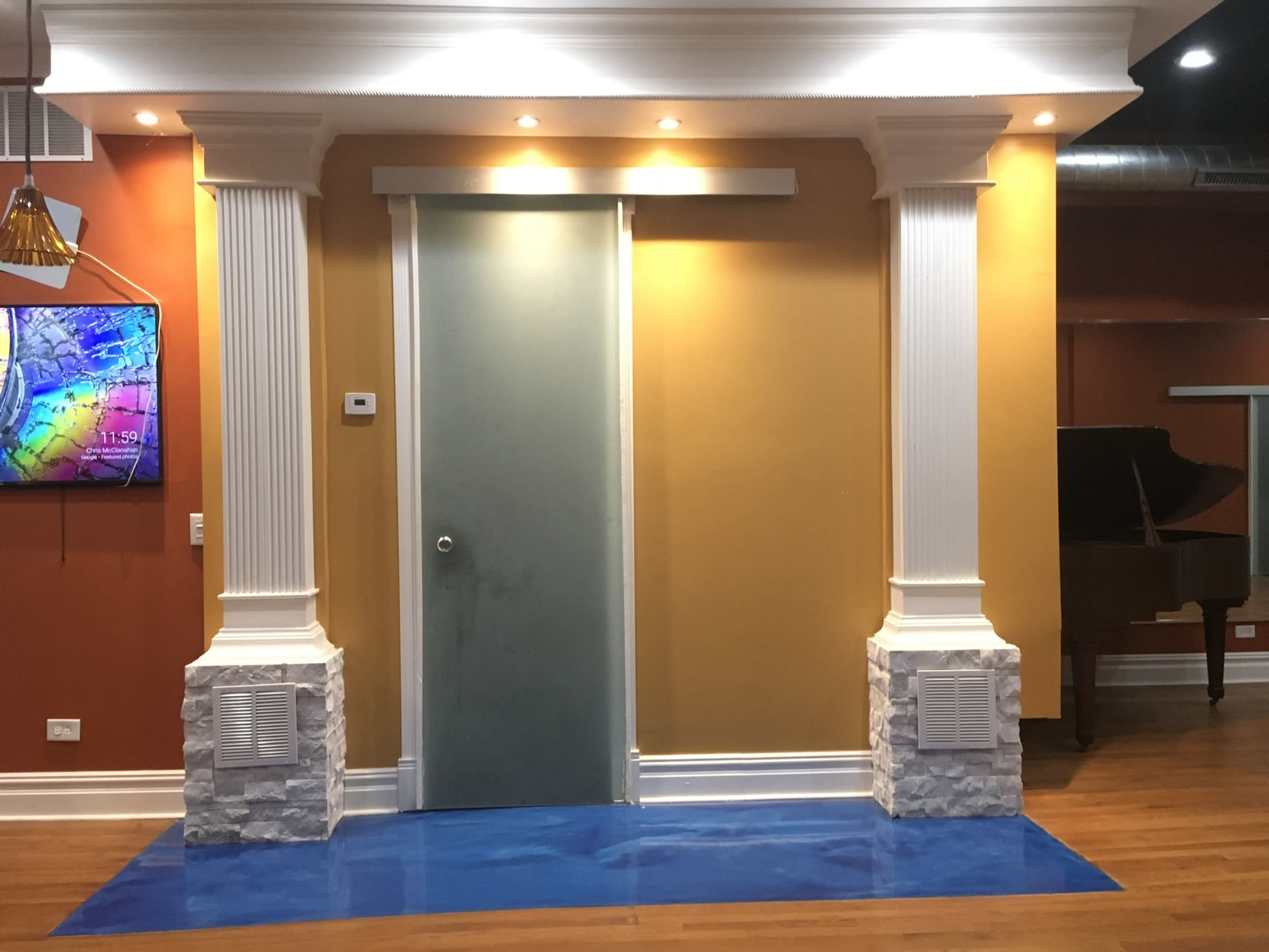A room featuring a glass sliding door between two decorative columns, with a television on the left and a piano in the background.
