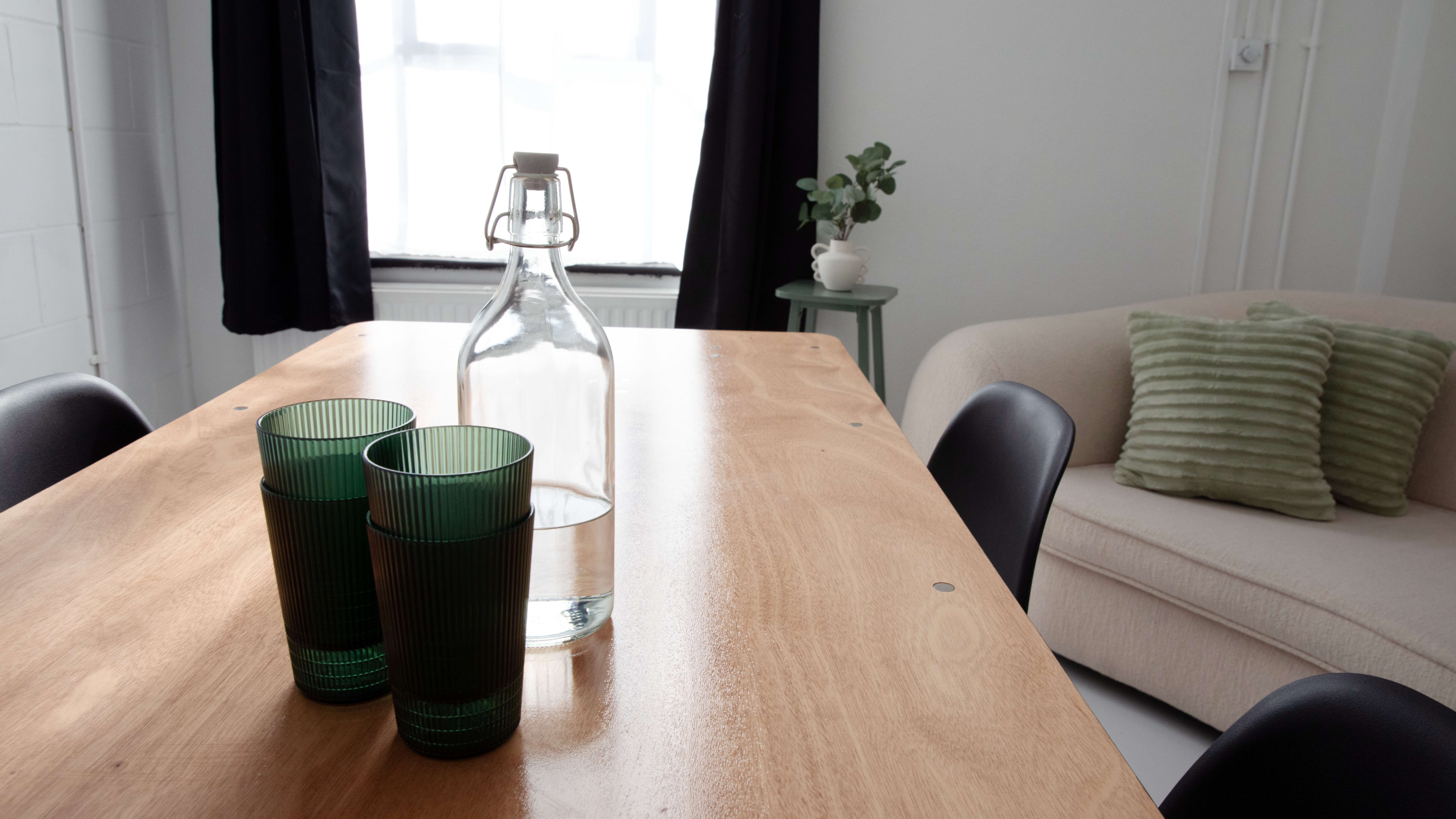 A wooden table with two green glasses and a bottle of water sits in front of a cream-colored sofa and a potted plant in a well-lit room.