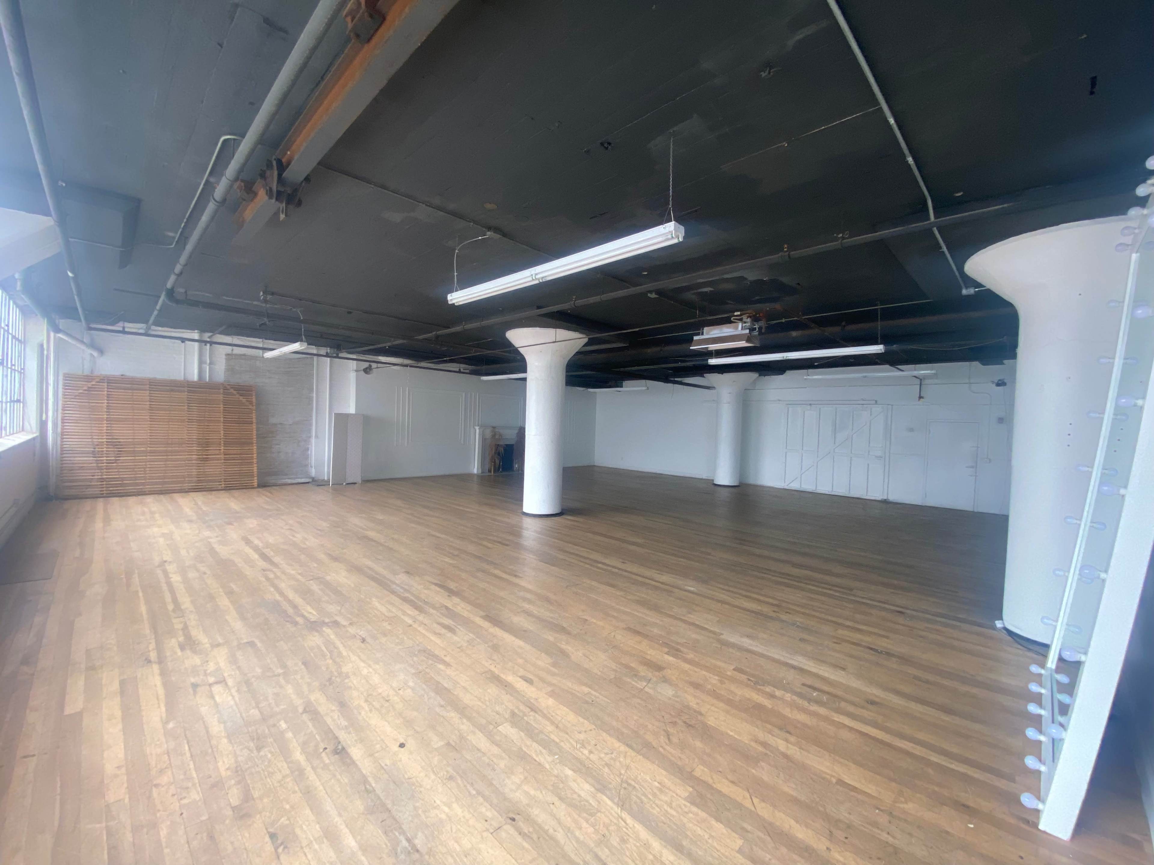 An empty industrial space with wooden floors and white pillars, illuminated by overhead fluorescent lights.