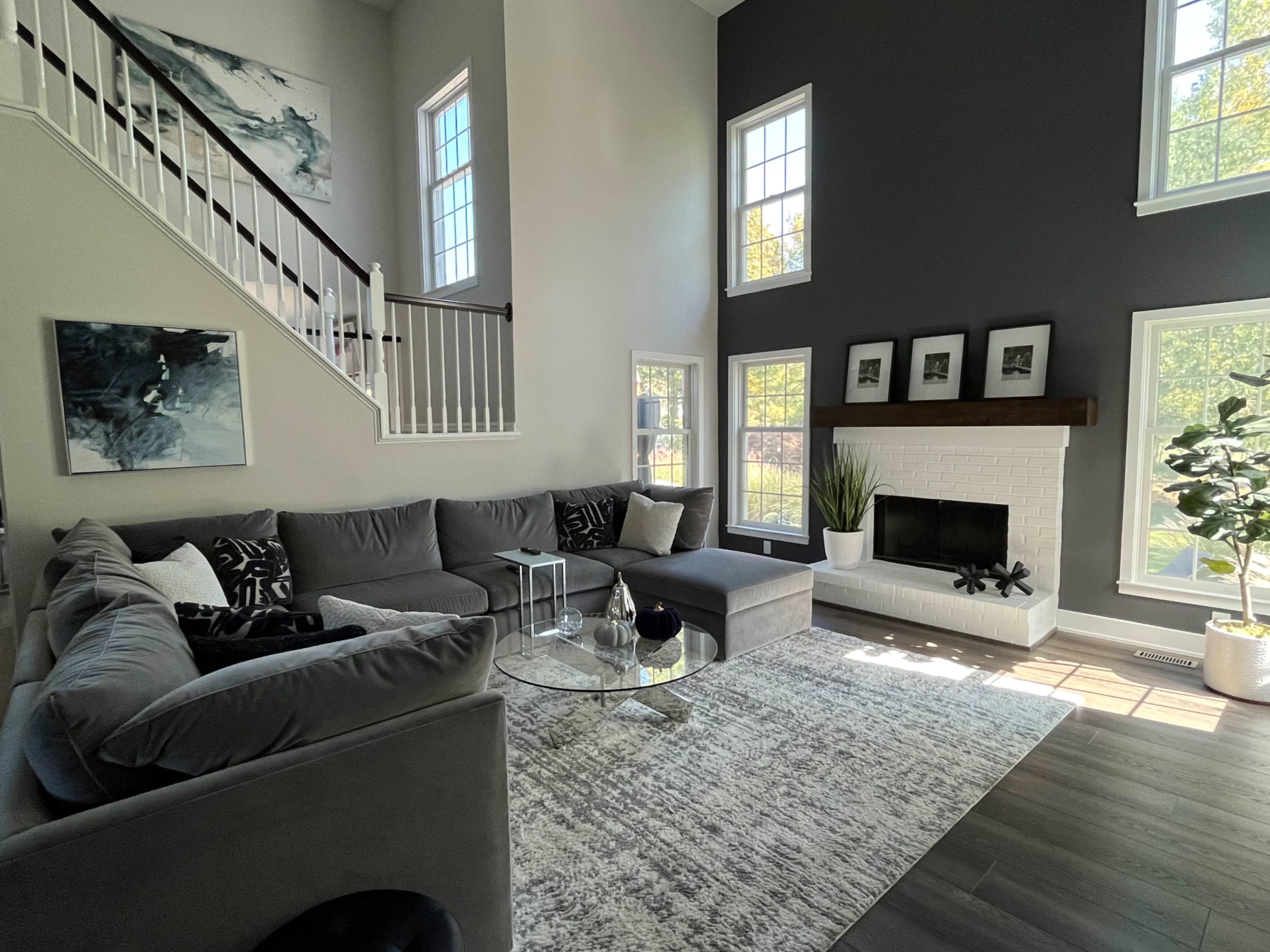 The living room features a large sectional sofa, a round glass coffee table, and a white brick fireplace, with large windows allowing natural light to fill the space.