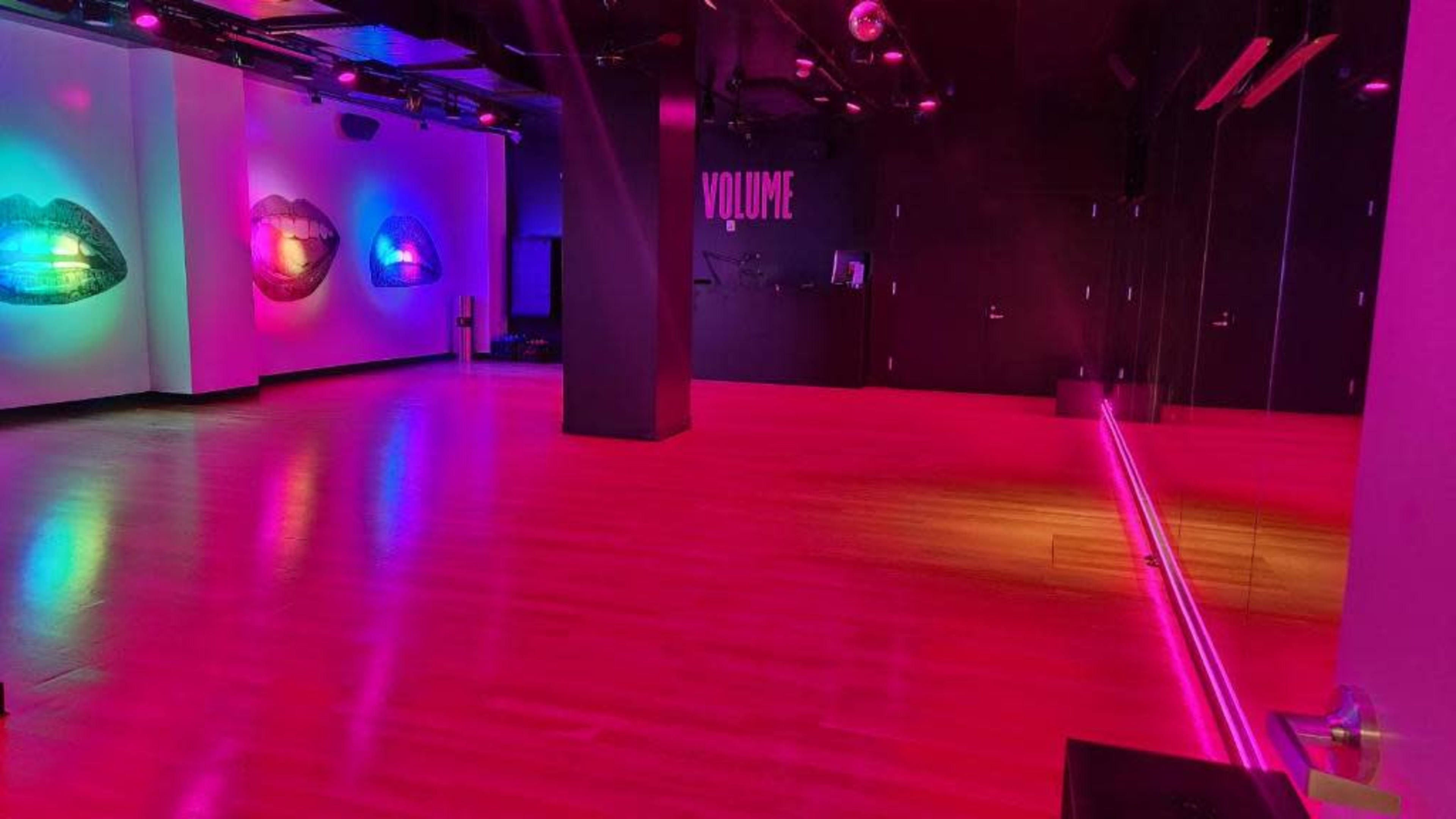 The image shows a dimly lit dance studio with colorful wall art and a mirrored wall, featuring the word "VOLUME" on one side.