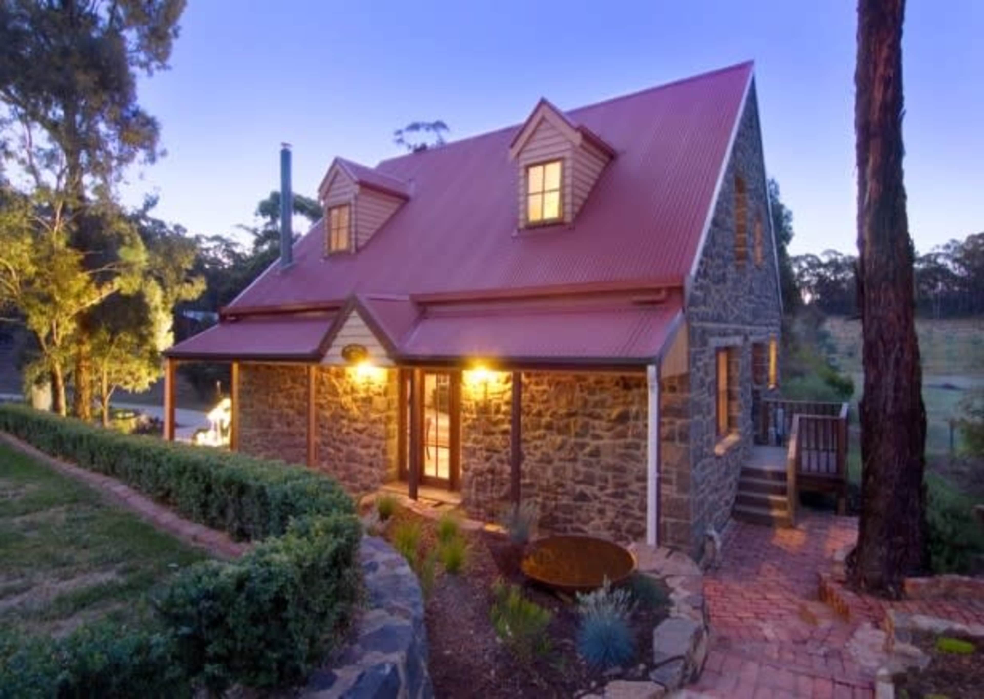 A stone cottage with a red roof is illuminated by warm light and surrounded by greenery at dusk.