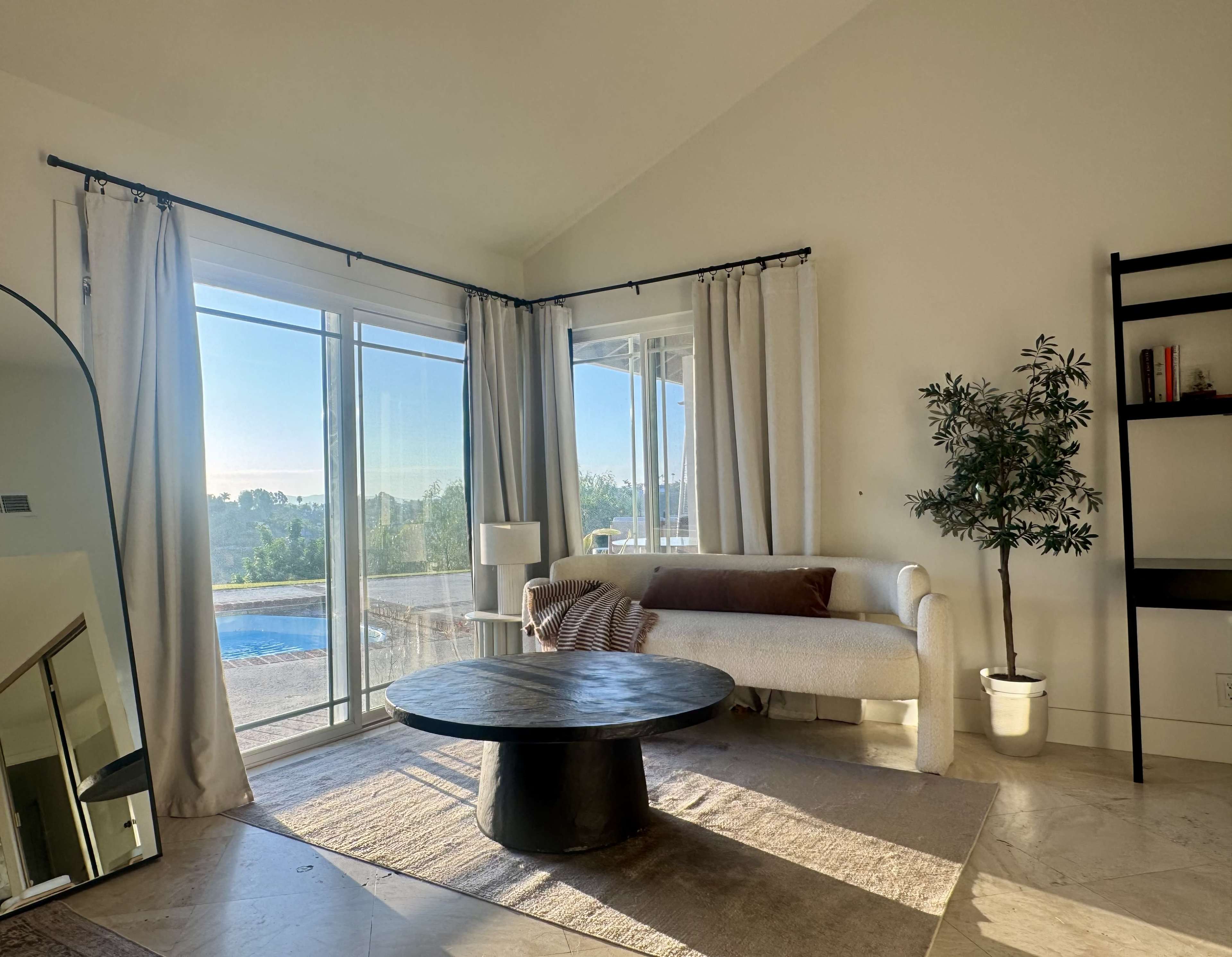 A bright living room features a white sofa, a round wooden coffee table, and large windows overlooking a pool area, with light-colored curtains and a potted plant in the corner.
