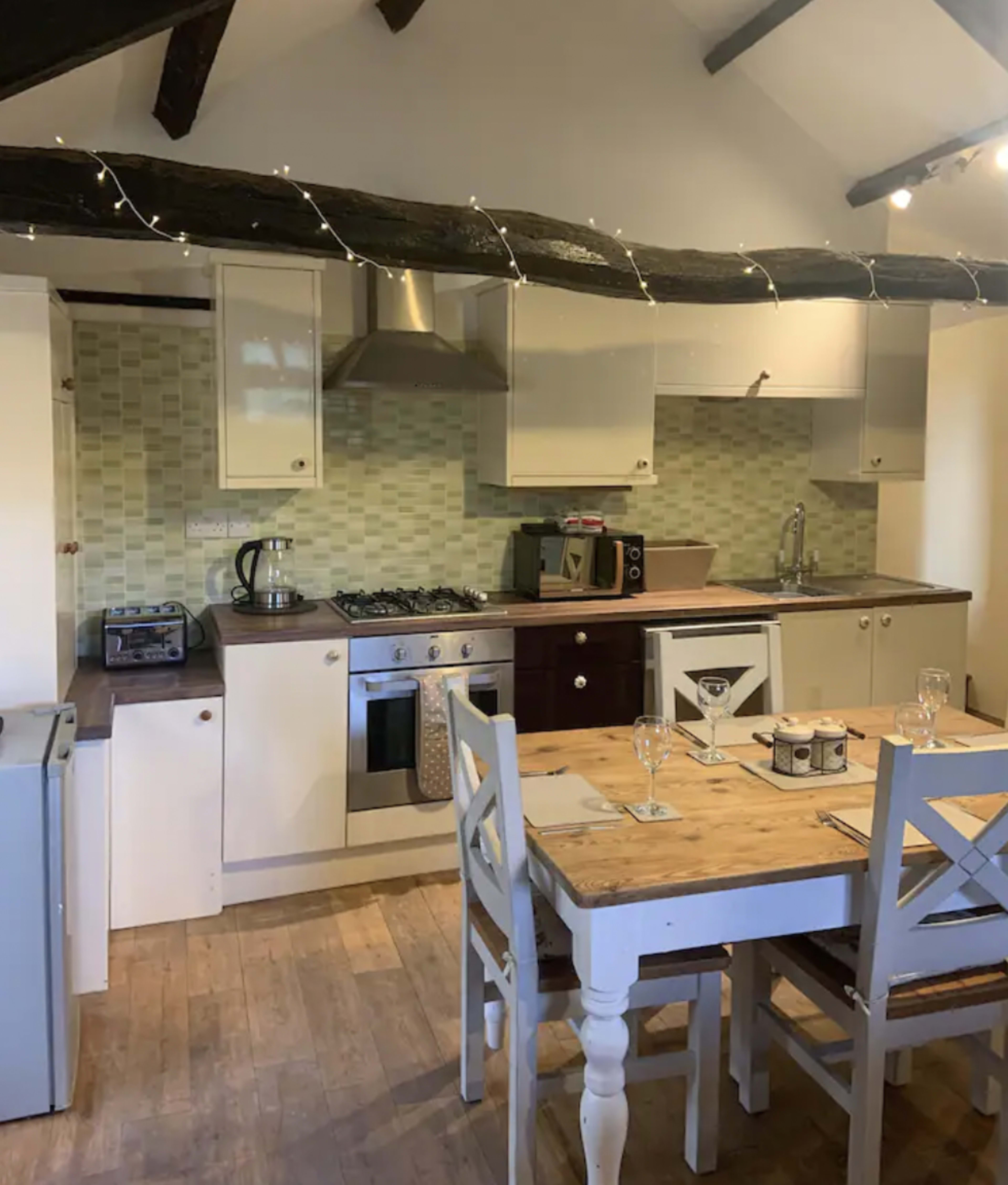 The image shows a modern kitchen with a gas stove, sink, and refrigerator, featuring wooden flooring and a dining table with four chairs.