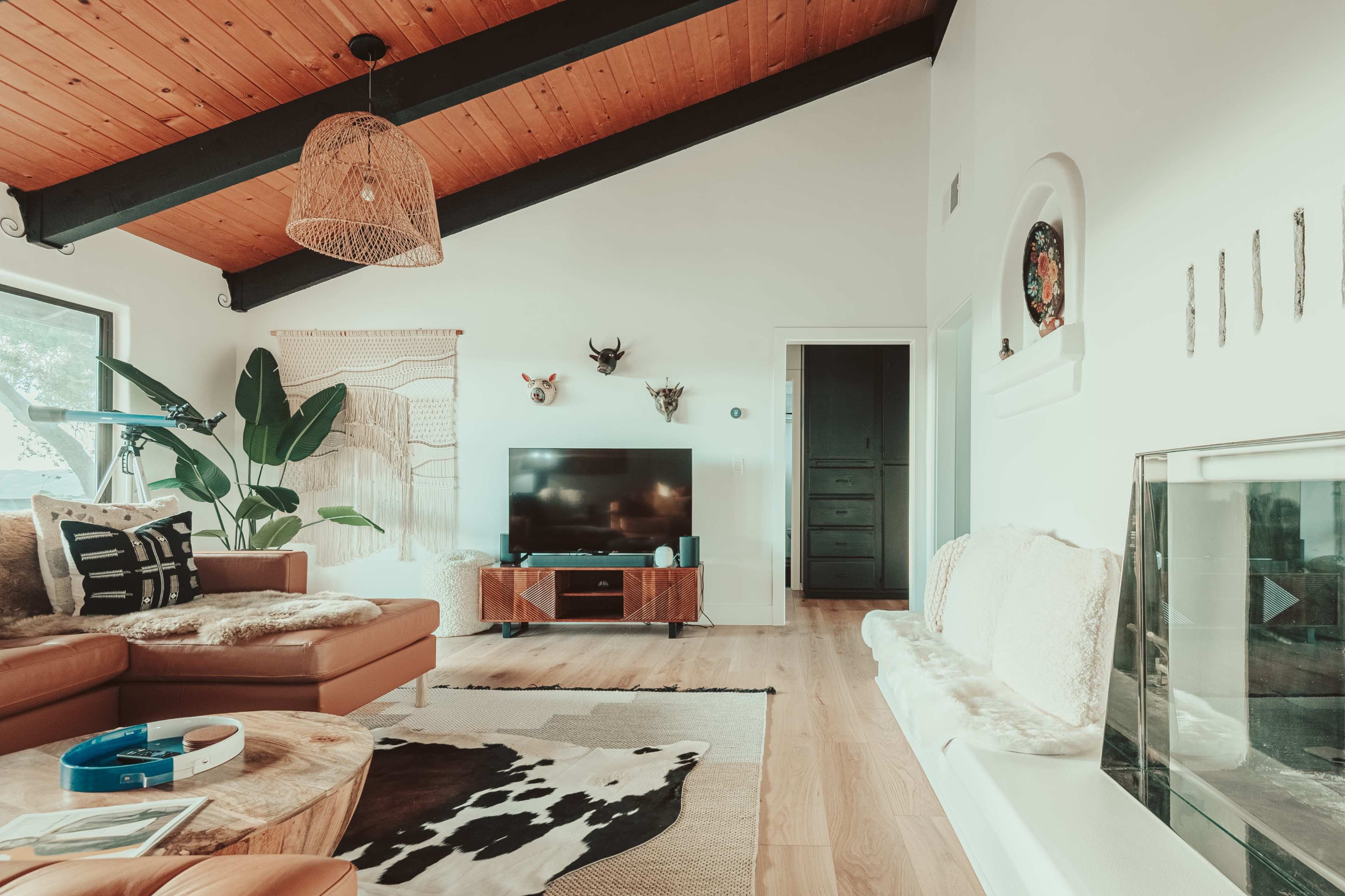 The living room features a brown leather sofa, a wooden coffee table on a cowhide rug, a television stand, and large windows with plants in a bright, airy space.