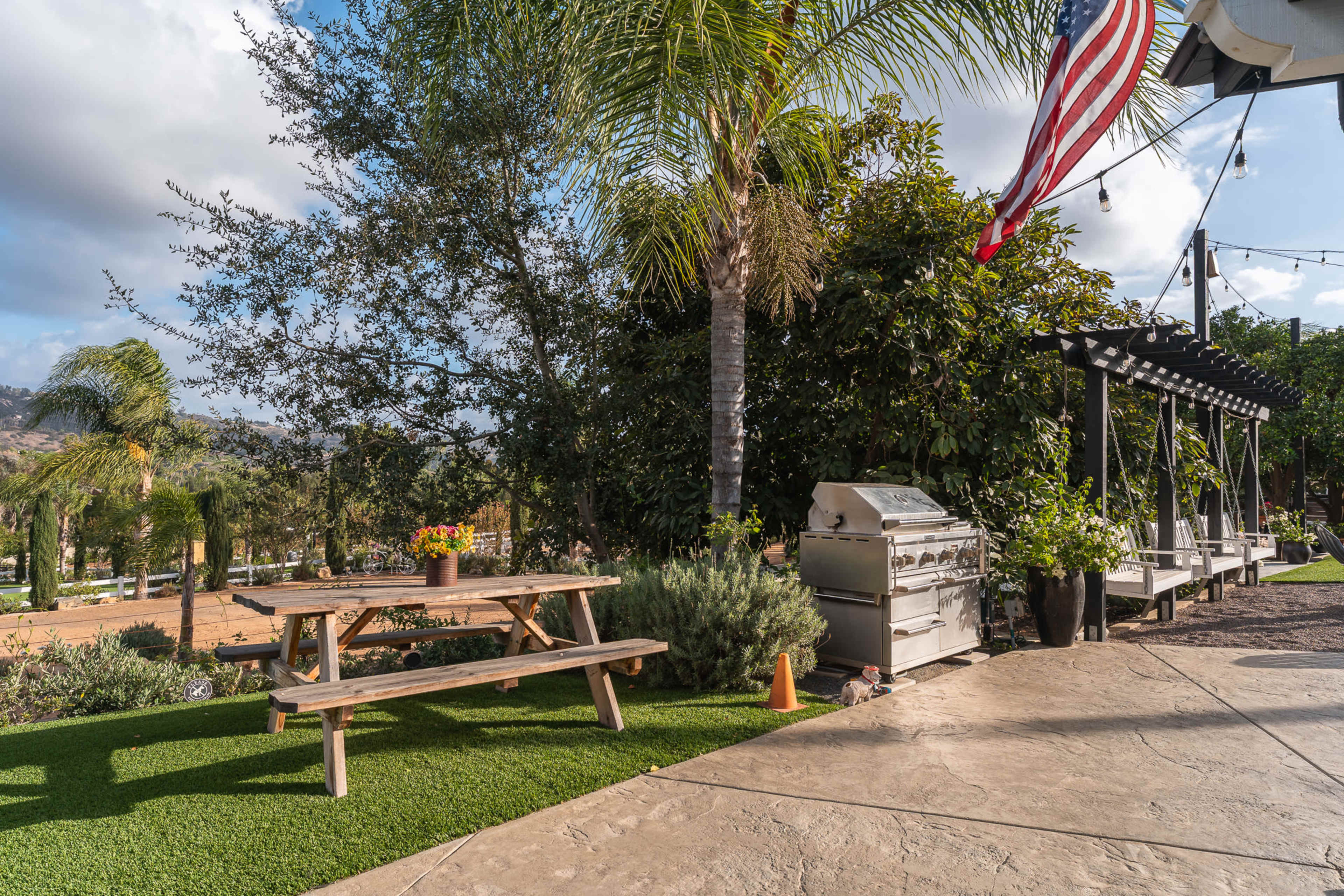 A picnic table sits on a grassy area next to a barbecue grill, surrounded by palm trees and plants, with a flag hanging nearby.