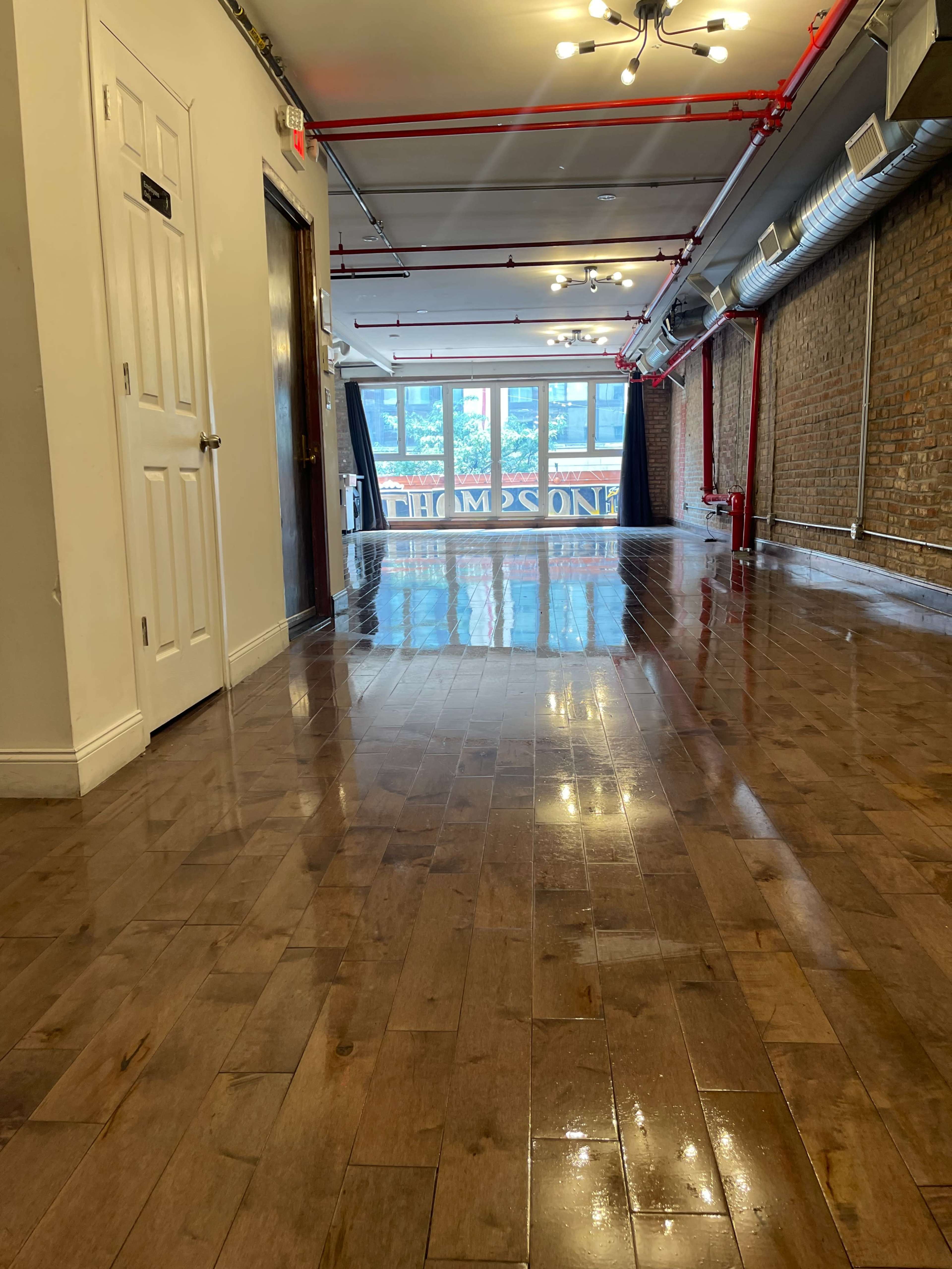 A long hallway with shiny wooden floors, white walls, and large windows at the far end, revealing an outdoor view.