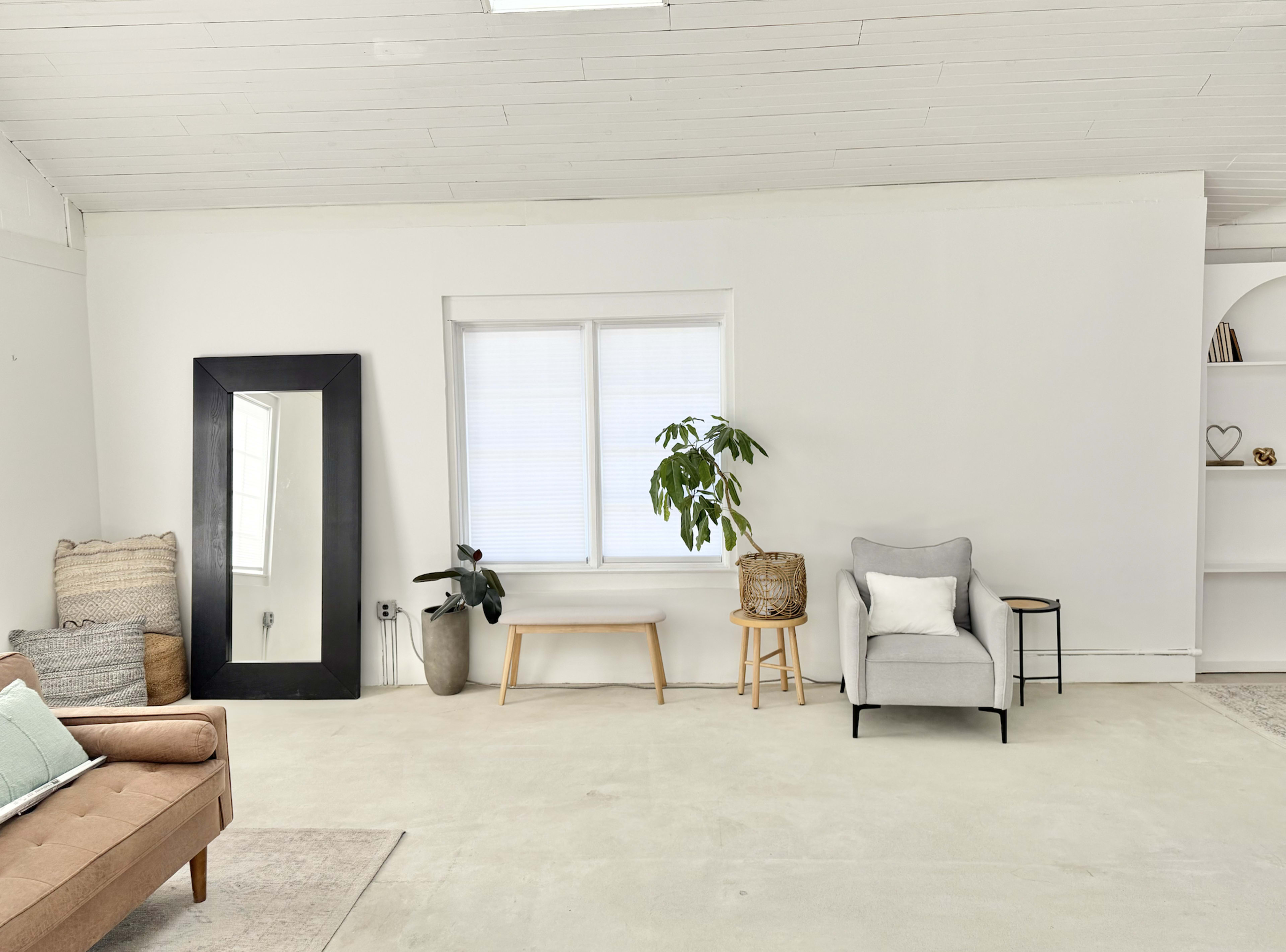 The image shows a bright, minimalist room featuring a large mirror, a plant, a small chair, and a couch.