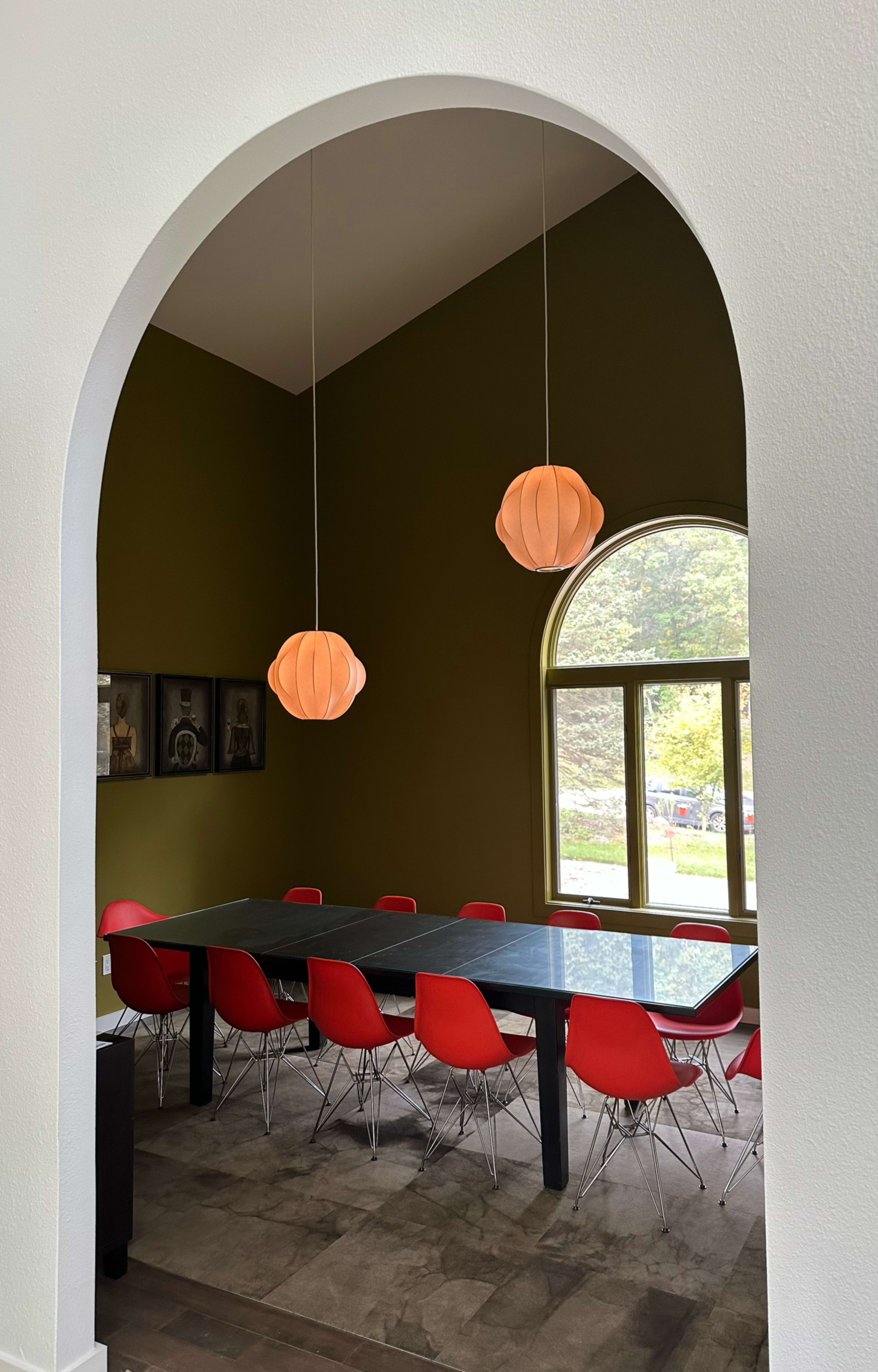 A dining room features a long black table surrounded by red chairs, illuminated by two pendant lamps with a soft, rounded design.