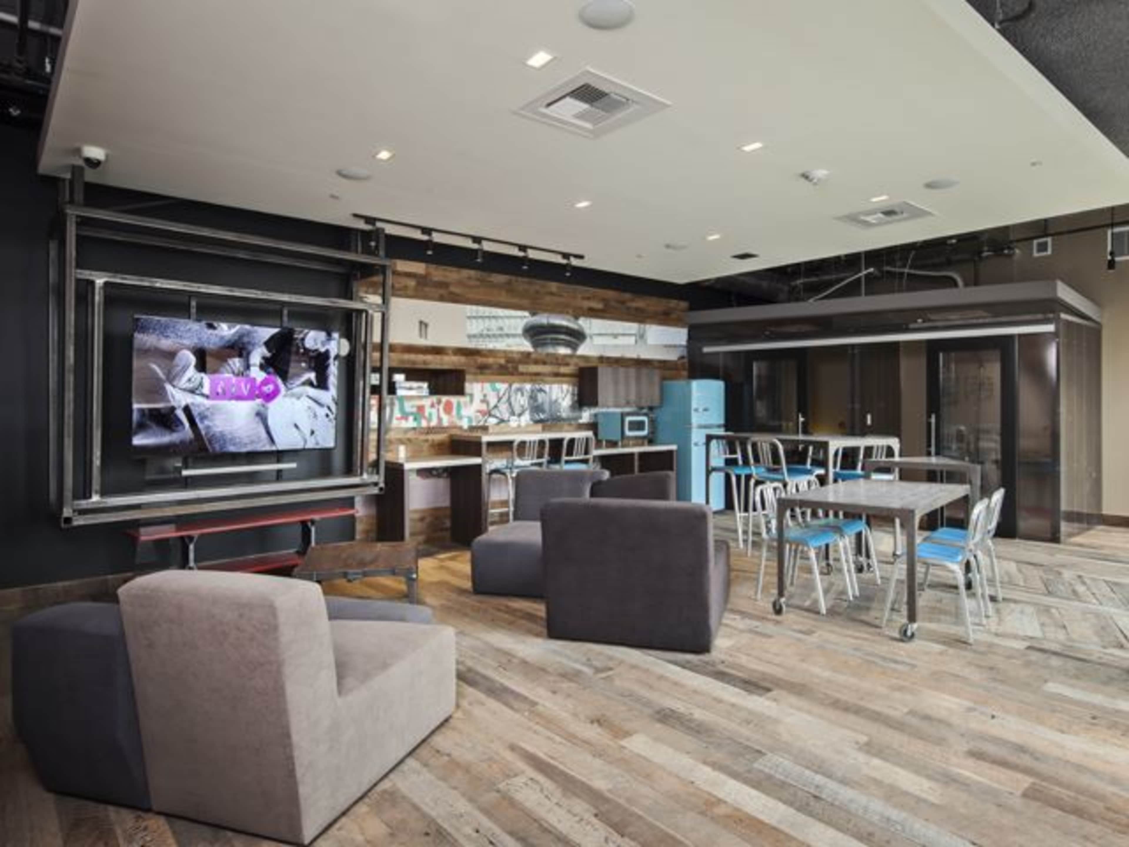 A modern communal room with a kitchen area, dining tables, and a large television mounted on the wall.