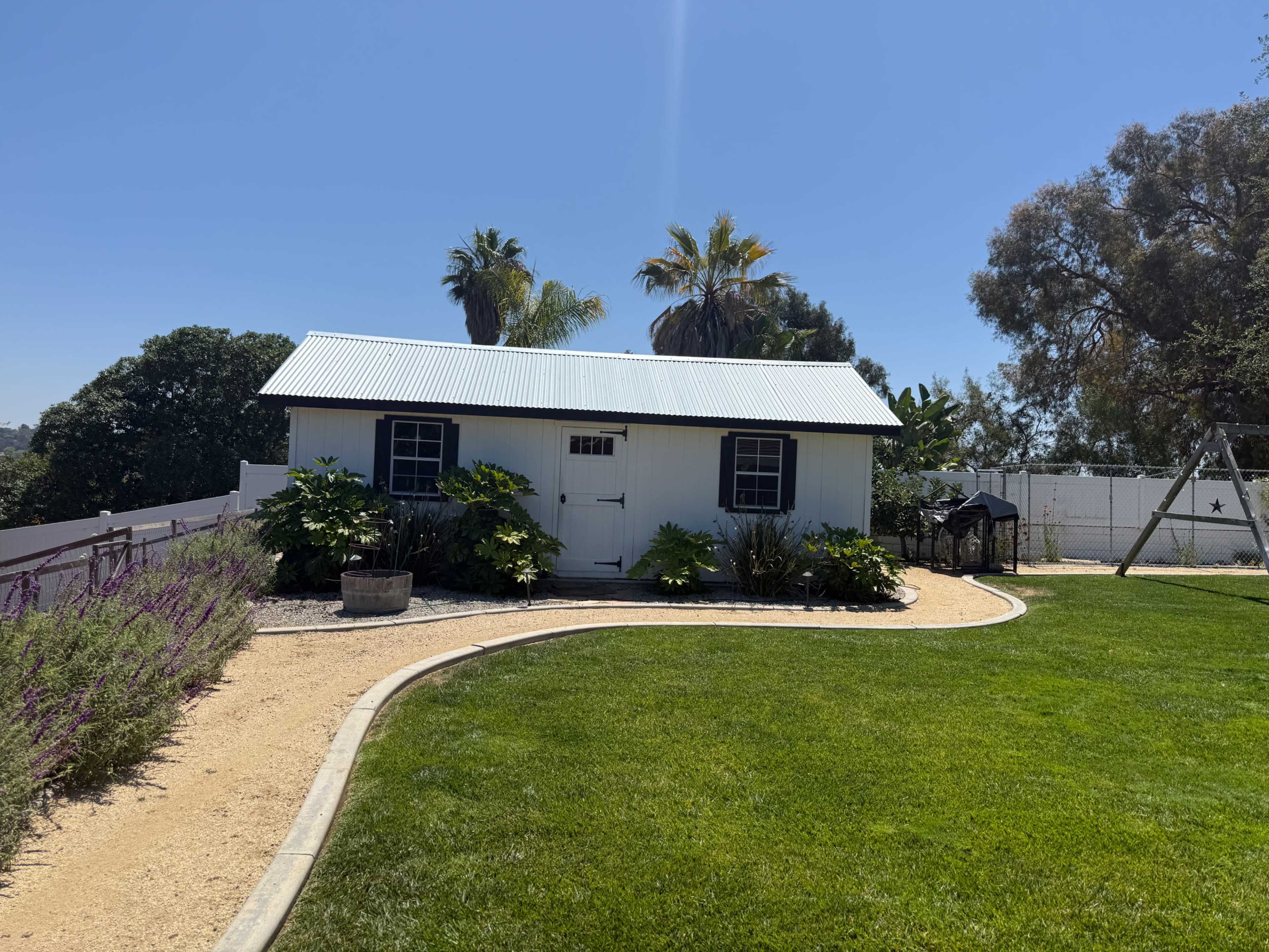 The image shows a white shed with a metal roof surrounded by green grass and landscaped plants.