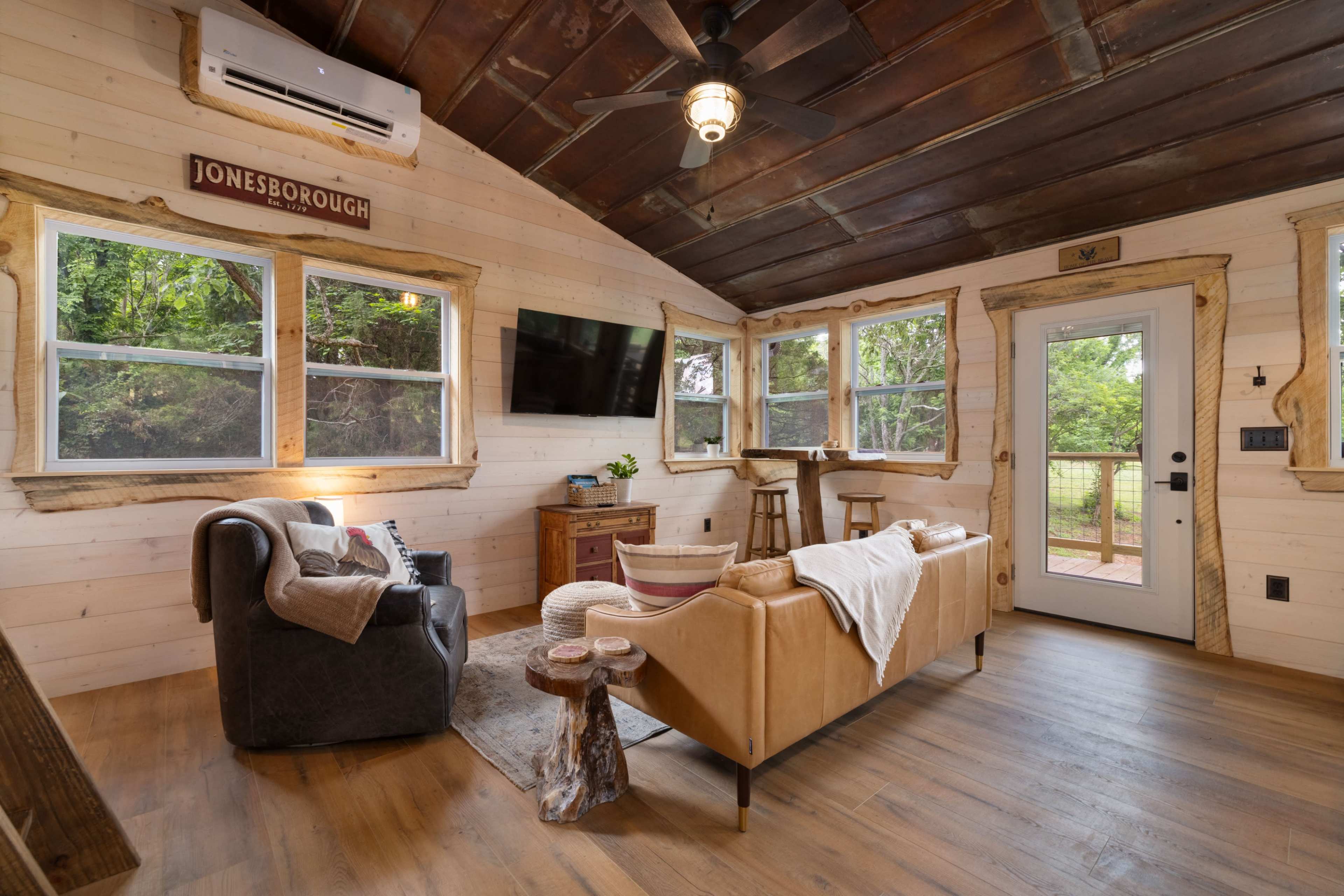 The image shows a cozy living space with wooden walls, a television mounted on the wall, a couch, and large windows providing natural light.