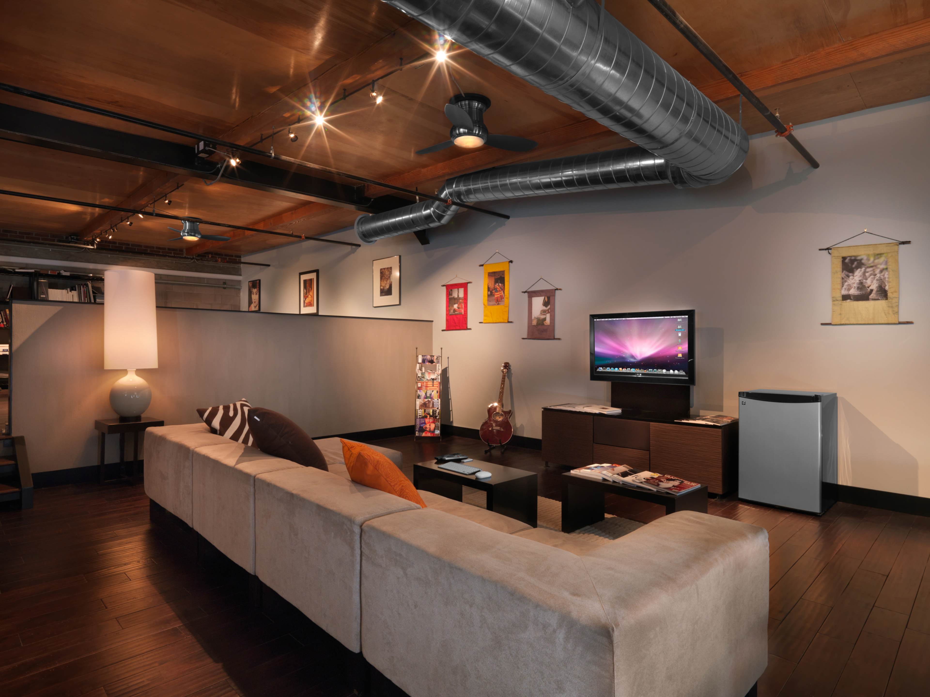 A modern living room features a beige sectional sofa, a television on a low table, and a small refrigerator, all in a space with wooden flooring and exposed metal ductwork above.