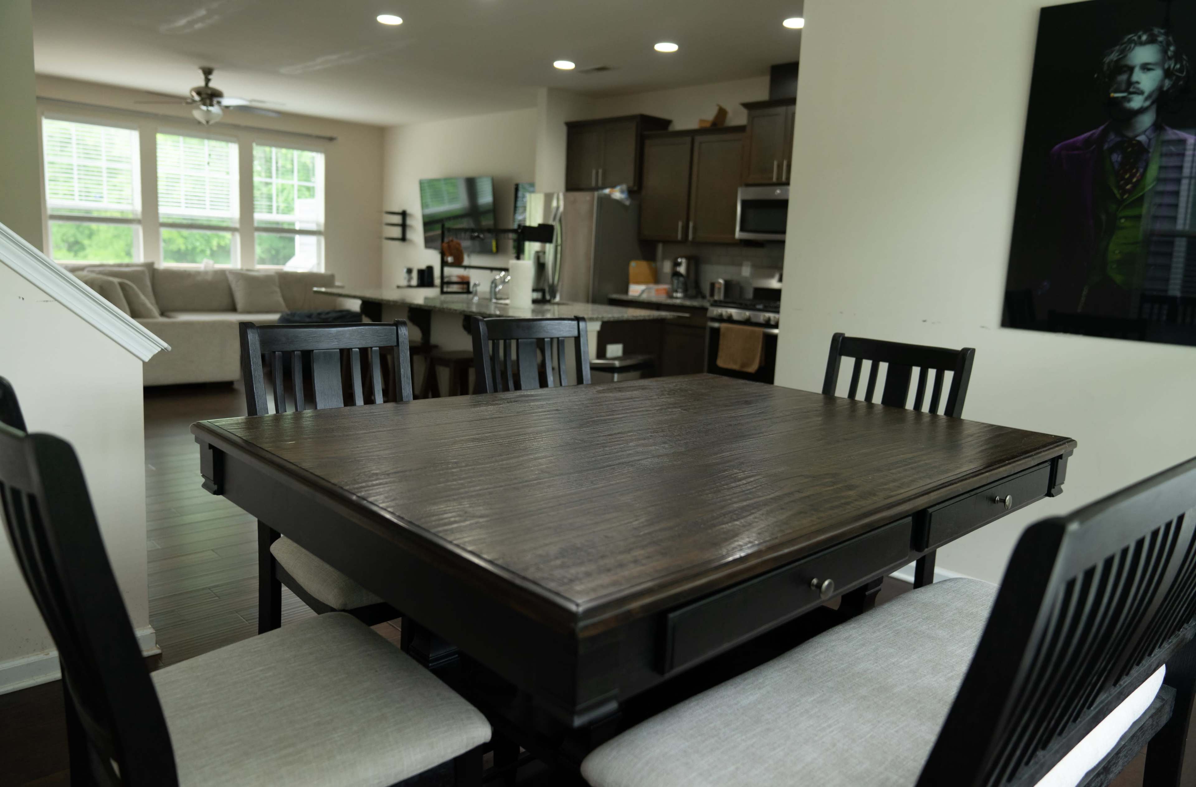 A dark wooden dining table surrounded by black chairs is set in an open-concept kitchen and living area with large windows.