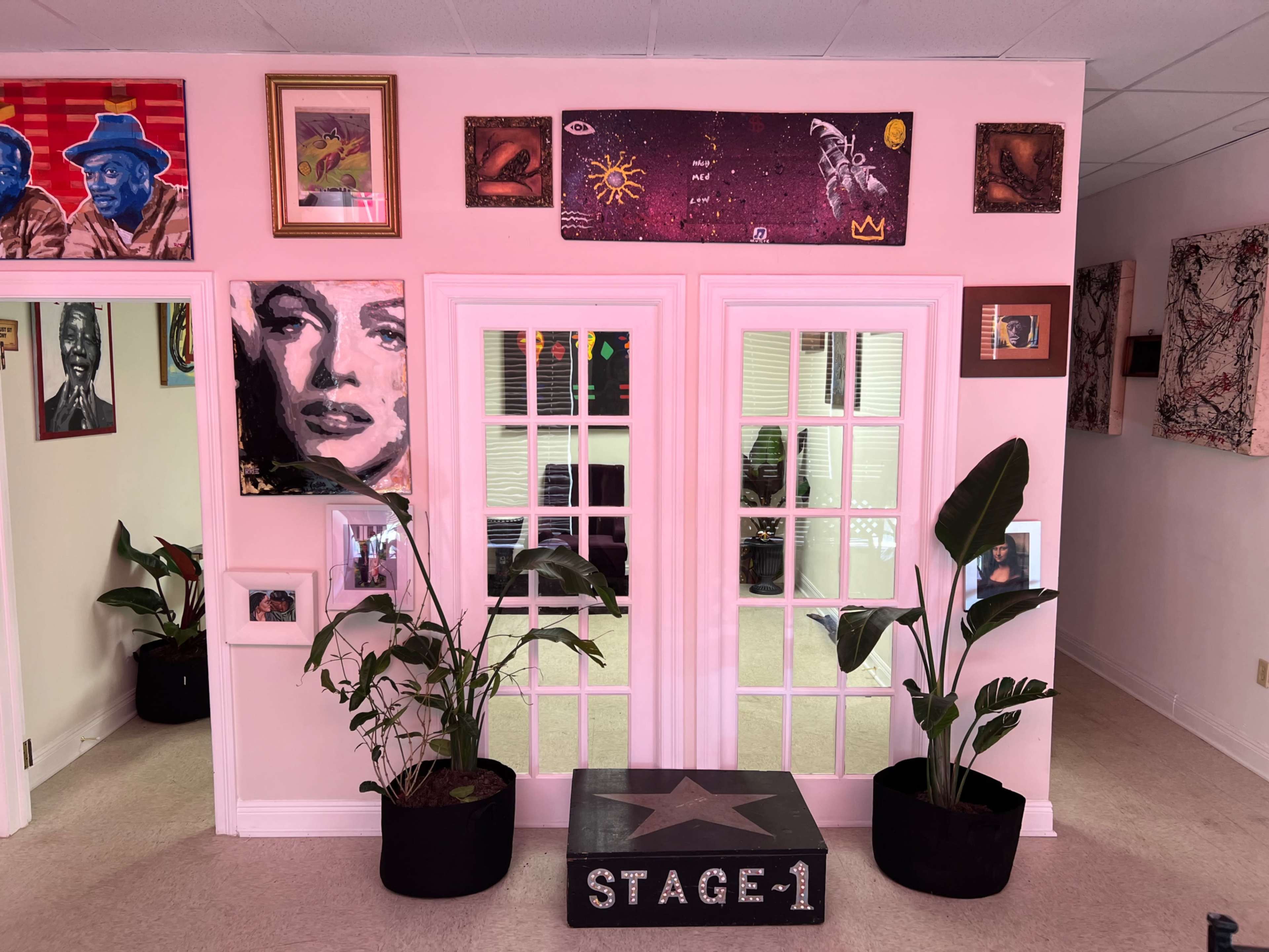 The image shows a doorway with glass-paneled double doors surrounded by various framed artworks and plants in two black pots.