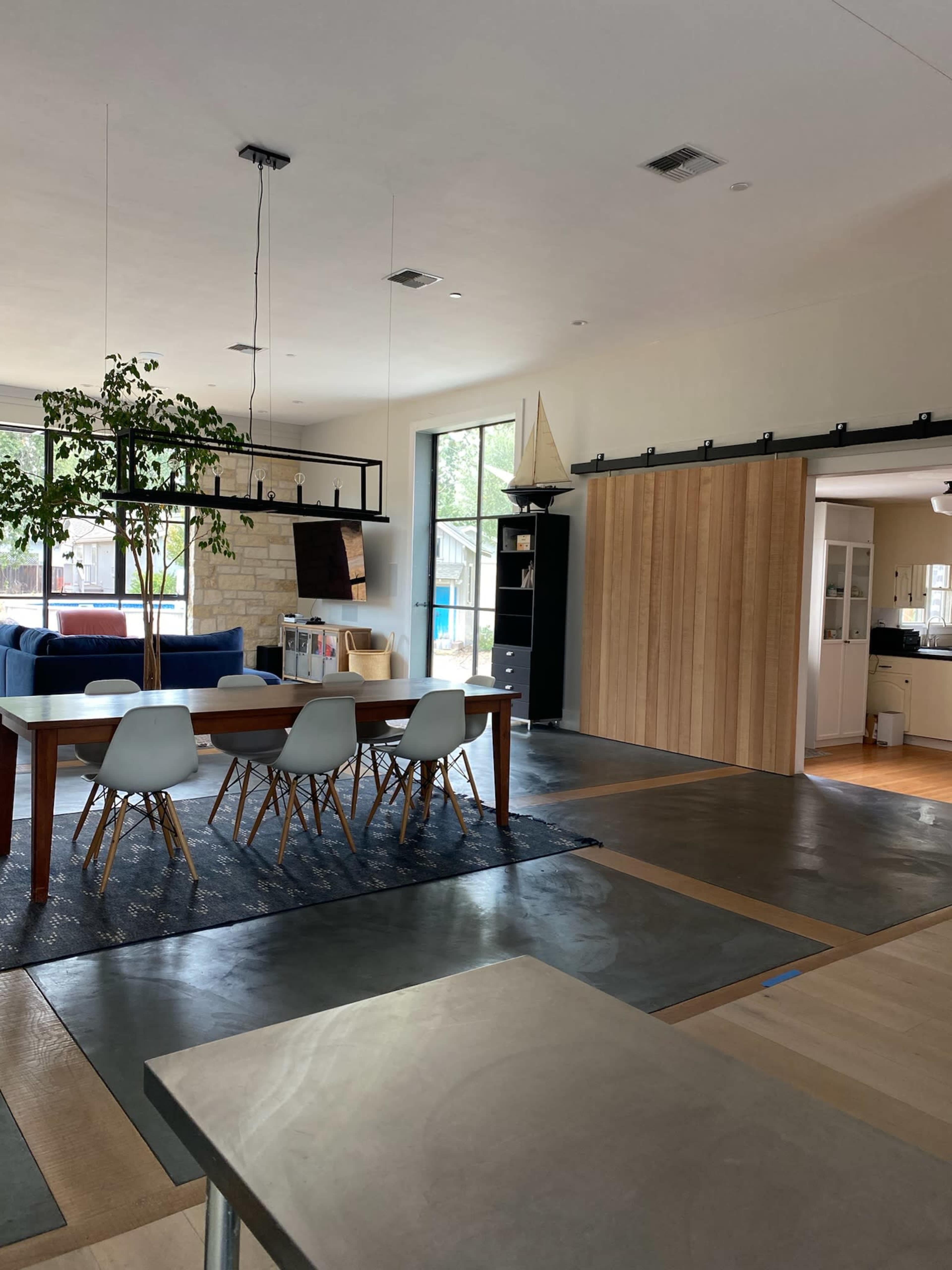 A modern interior space with a dining area featuring a wooden table and white chairs, a living area with a blue sofa, and a sliding barn door leading to a kitchen.