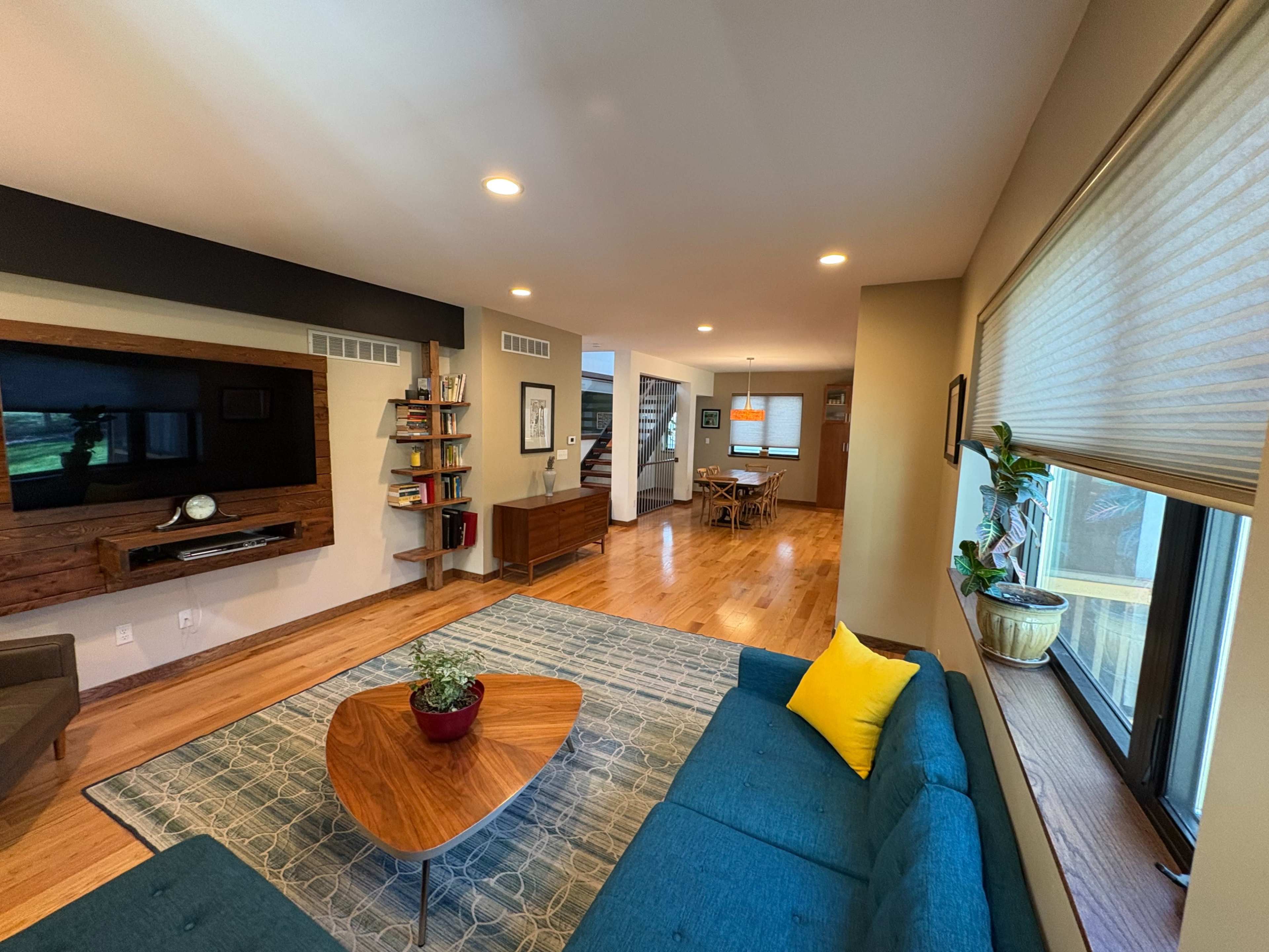 The image shows a spacious living room featuring a blue couch, a wooden coffee table, a television mounted on the wall, and a dining area in the background.