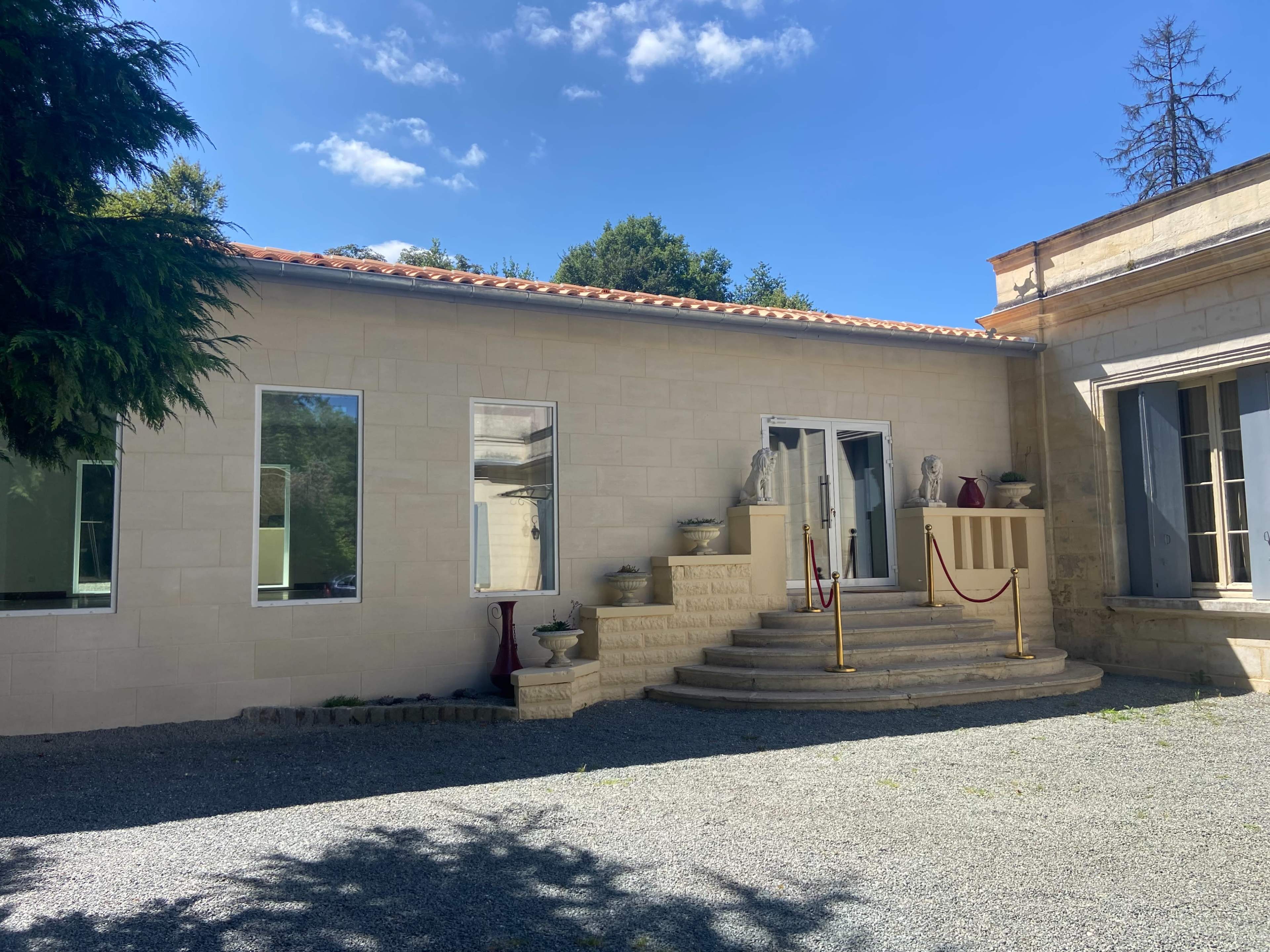 The image shows a beige stone building with steps leading up to a set of double doors, flanked by decorative statues and surrounded by a gravel area.