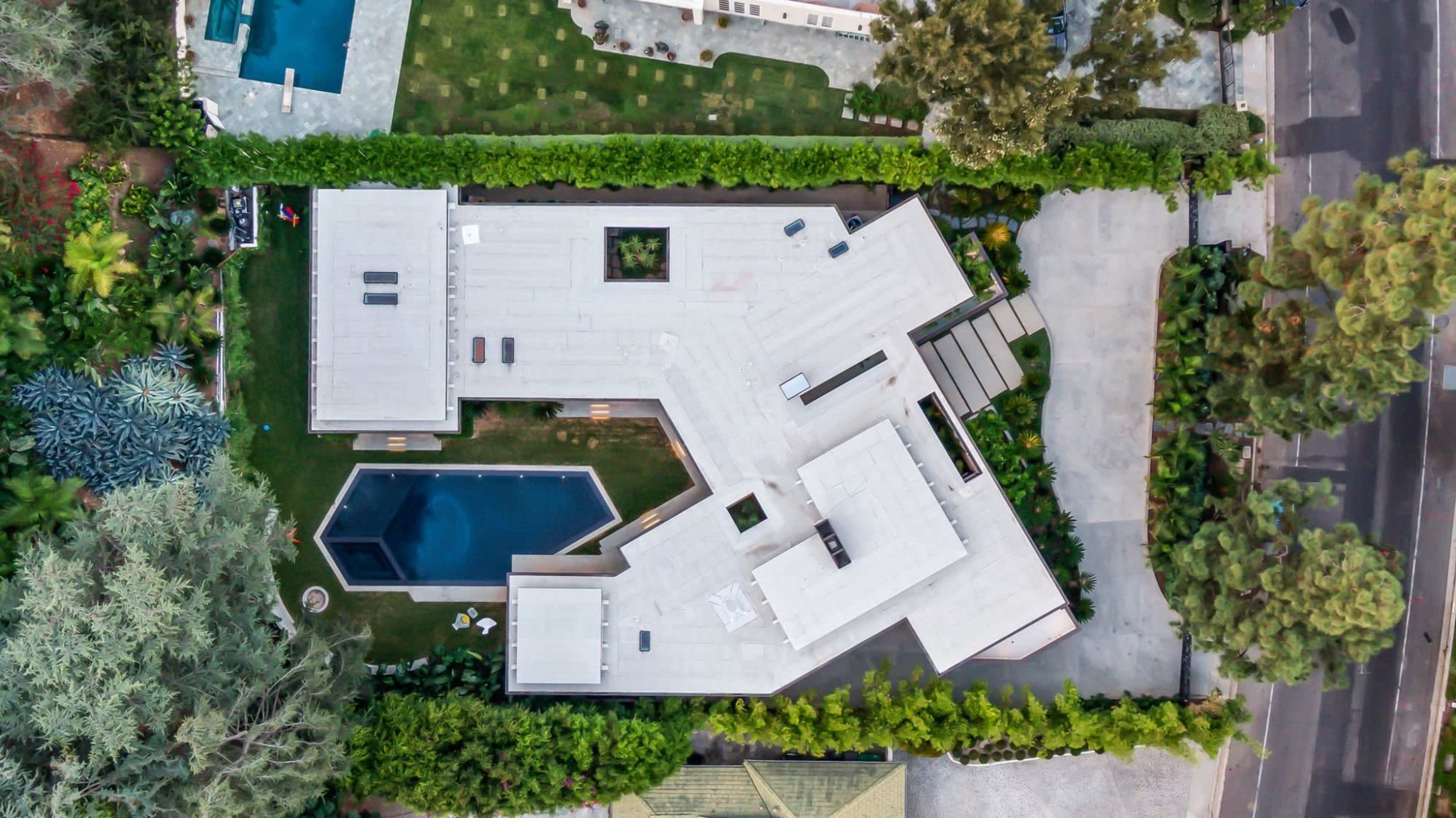 An aerial view shows a contemporary house with angular architecture, surrounded by greenery and featuring a swimming pool.