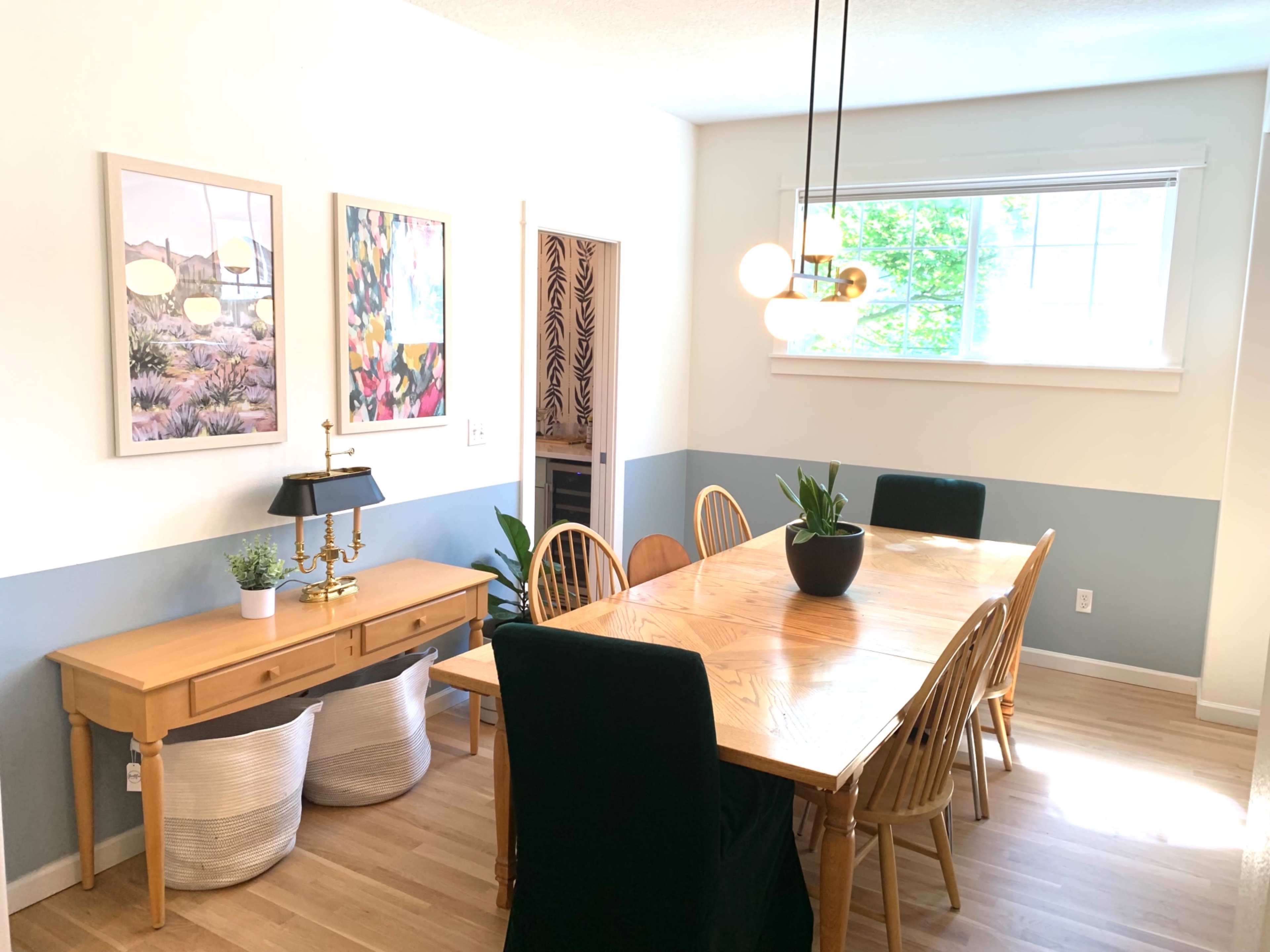 A dining room features a long wooden table surrounded by chairs, with artwork on the walls and a lamp on a console table.