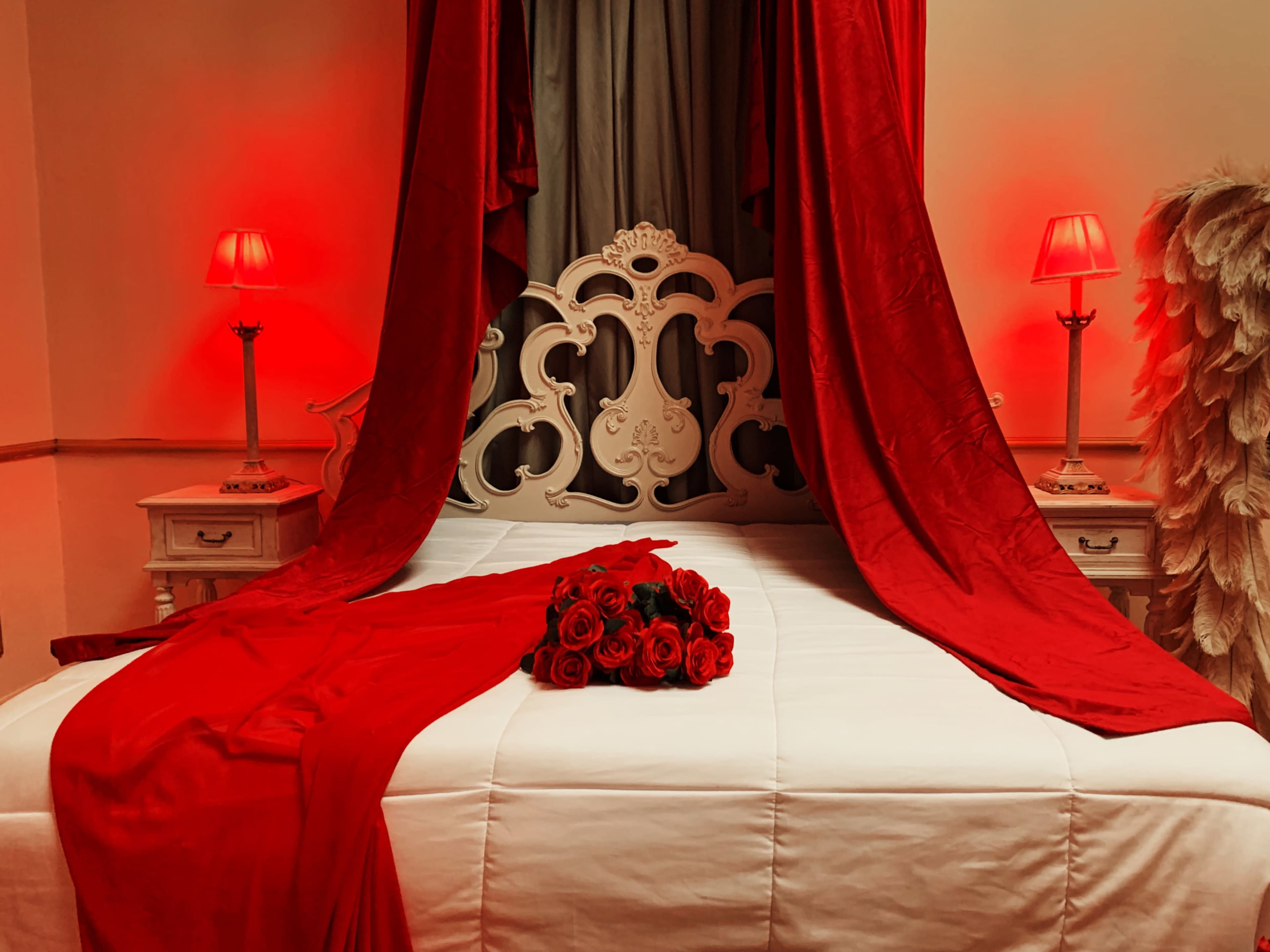 A neatly arranged bed with red drapery and lamps, adorned with a bouquet of red roses at its center.