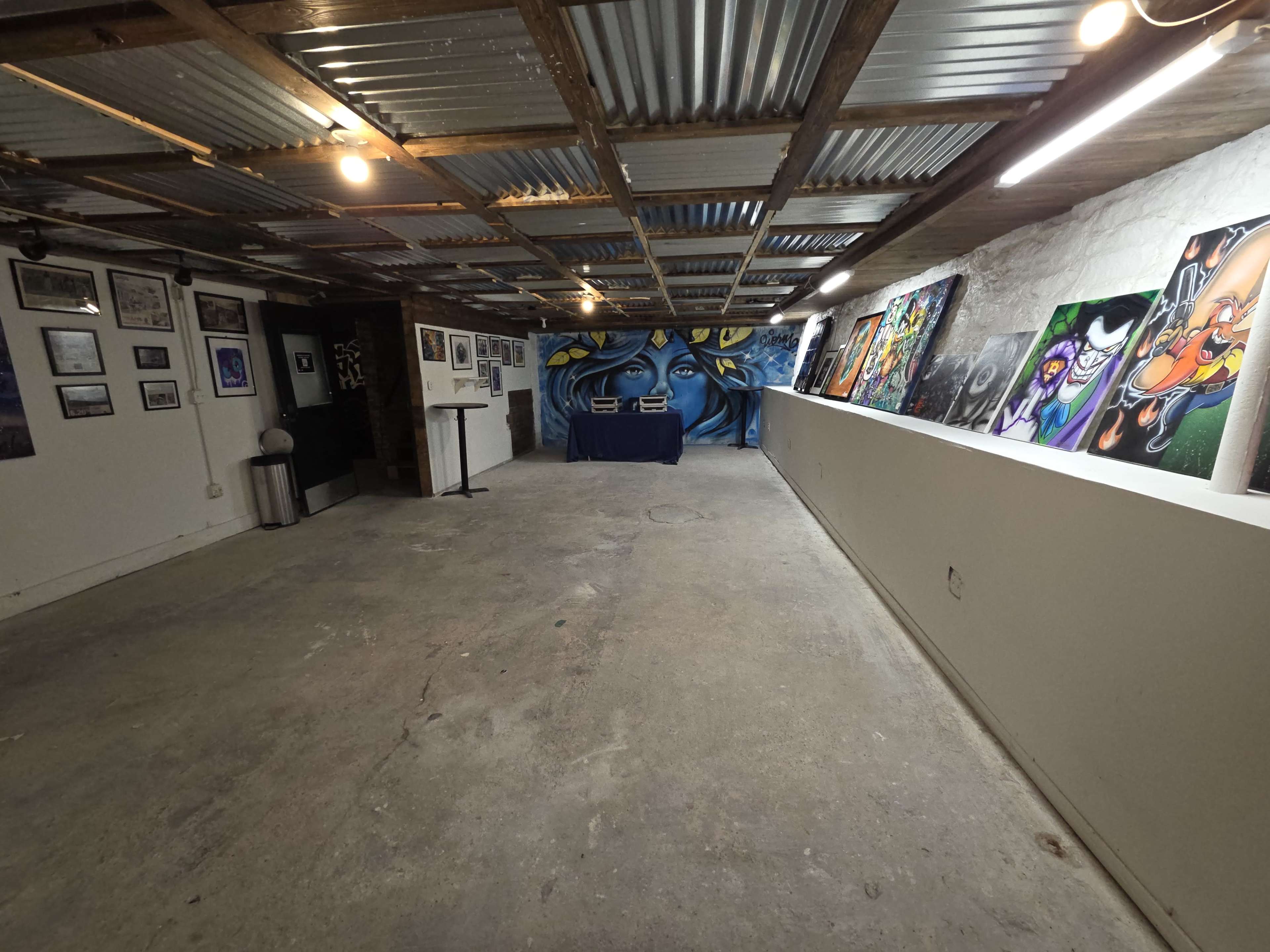 The image depicts a spacious gallery-like room with concrete flooring, metal ceiling panels, and a long wall displaying various artworks, while a table is placed in the center against a large mural at the far end.