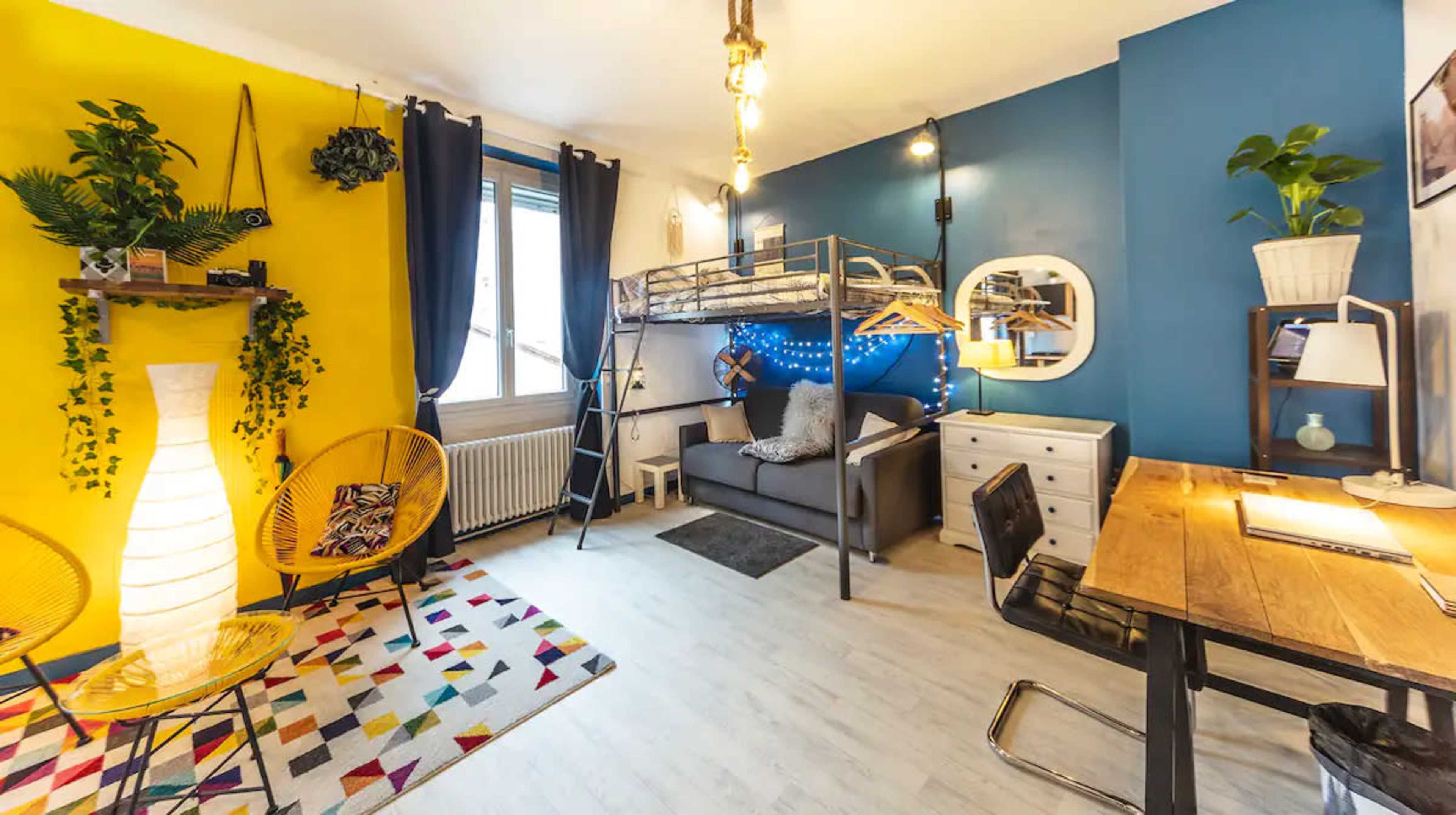 The image shows a colorful studio apartment featuring a lofted bed, a gray sofa, a desk with a chair, and decorative plants on the walls.