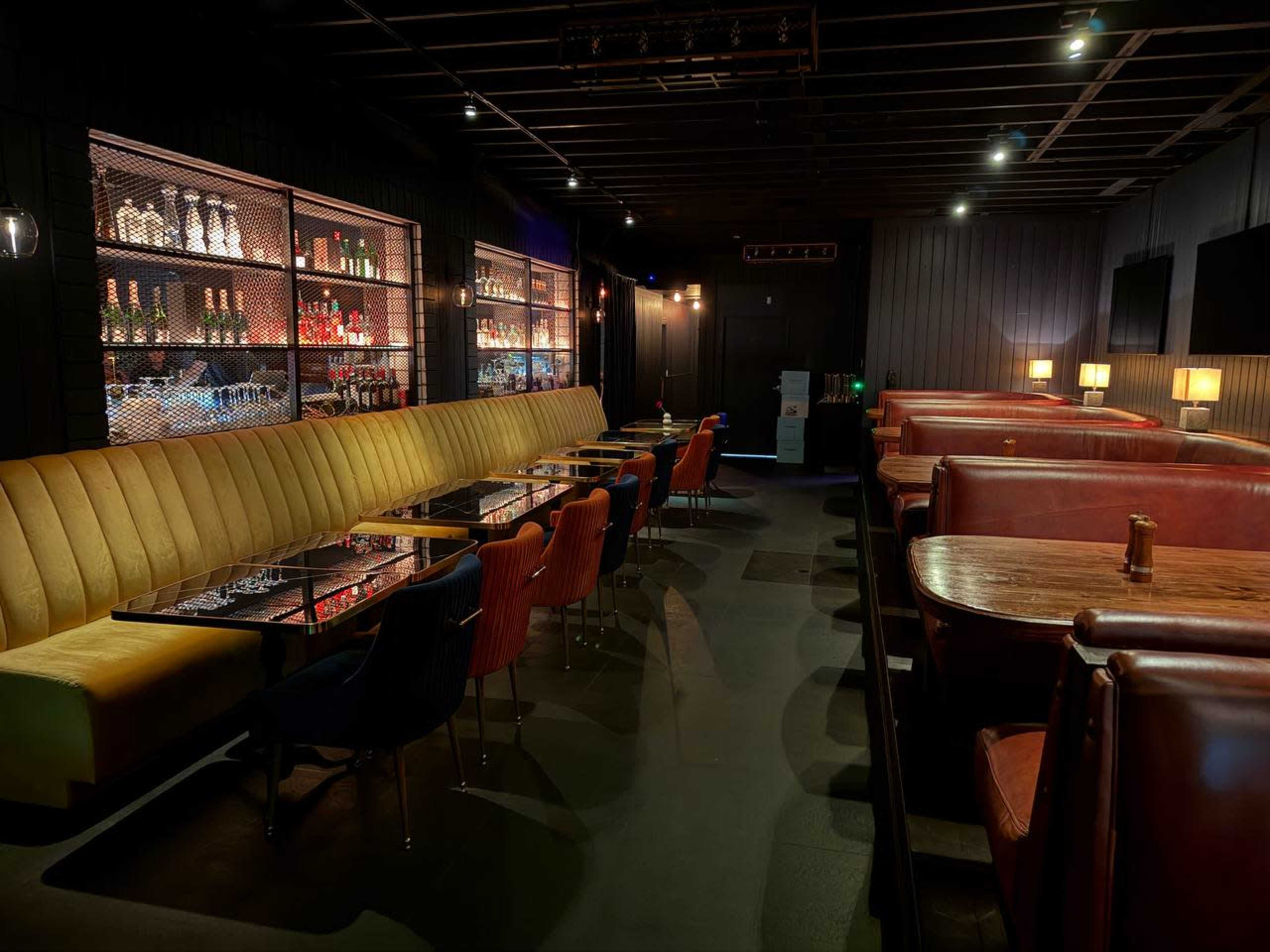 The image shows a modern restaurant interior with a mix of plush seating, wooden tables, and a well-stocked bar in the background.
