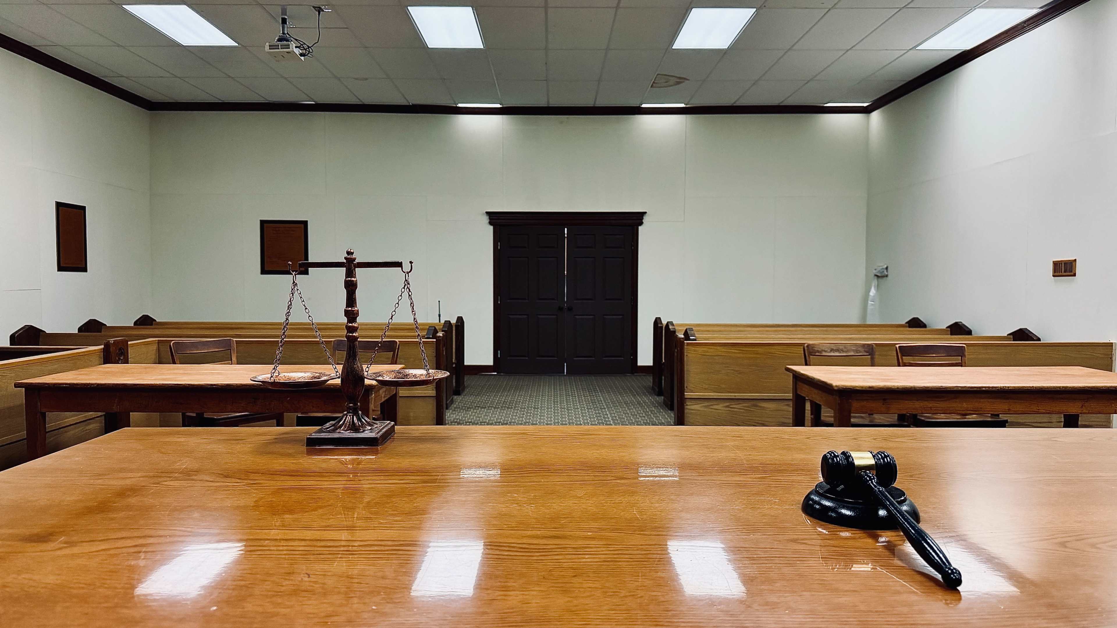 The image shows the interior of an empty courtroom with wooden benches, a gavel, and a scale of justice on a polished wooden table.