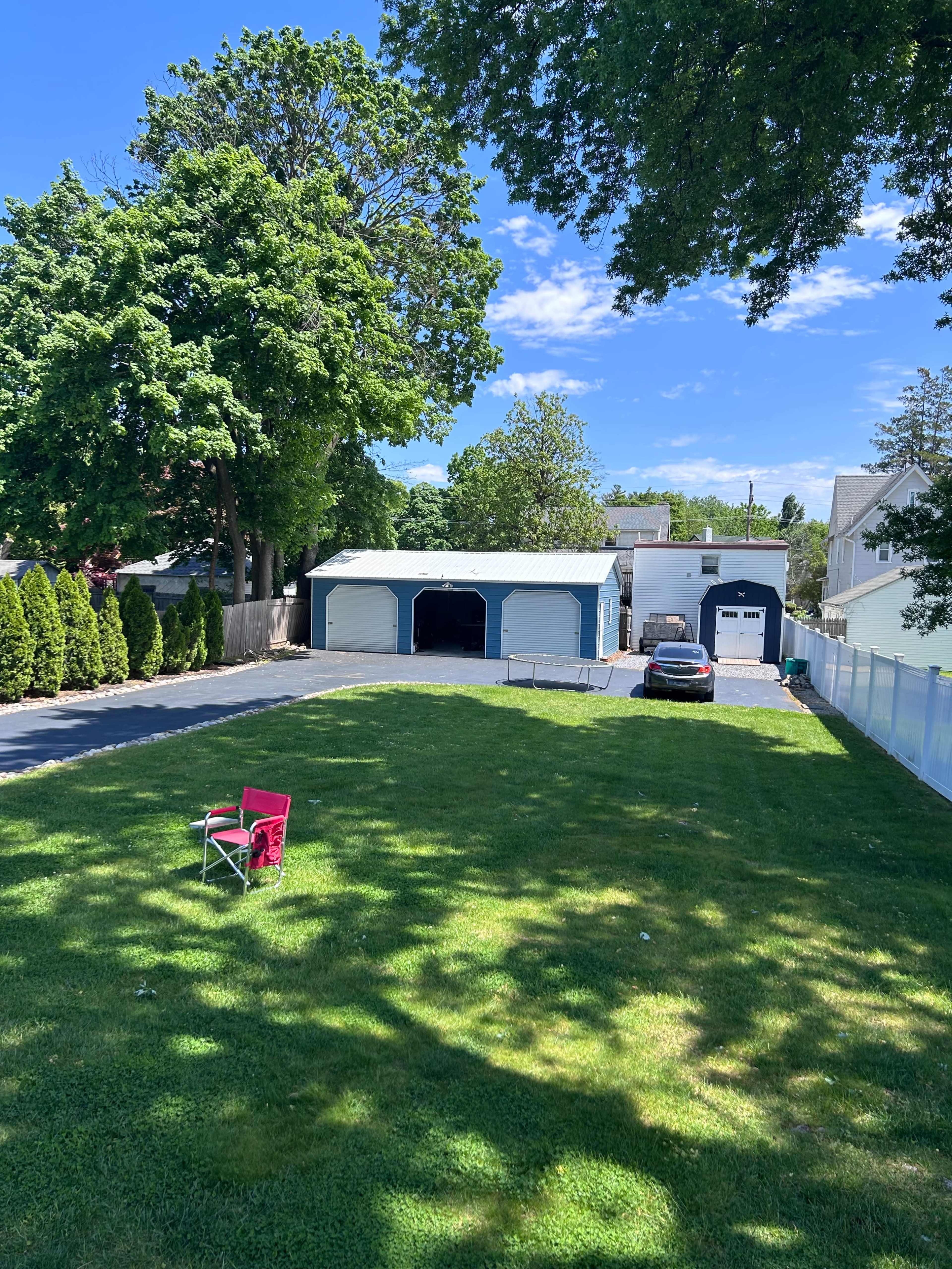 A grassy lawn is visible in the foreground with a single red chair, while a driveway leads to a two-car garage and a house in the background, framed by trees and blue sky.