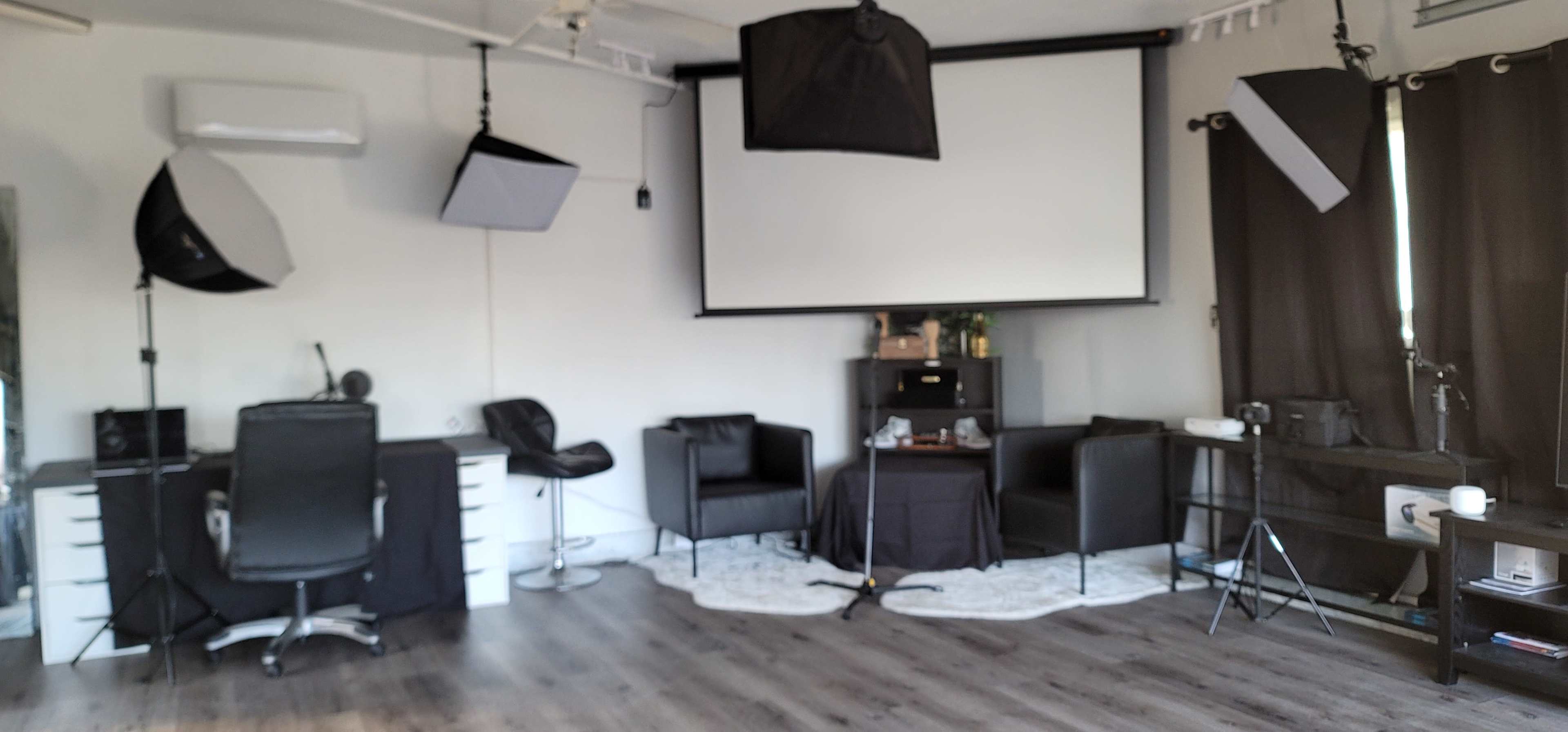 The image depicts a well-equipped studio space featuring a large projection screen, multiple lighting setups, and various black furniture pieces arranged around a central table.