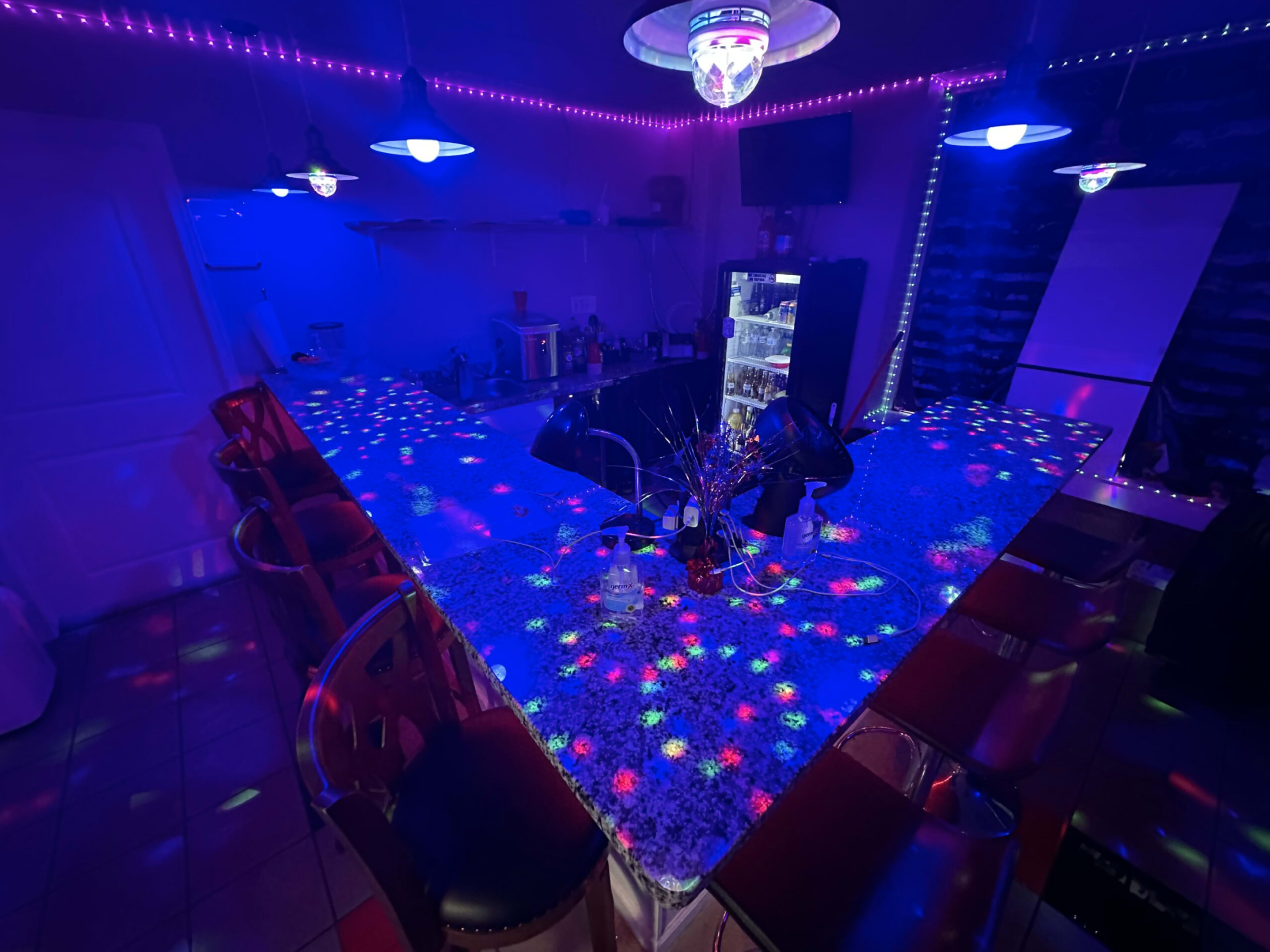 A dimly lit bar area features a brightly patterned countertop illuminated by colorful lights and a disco ball hanging from the ceiling.