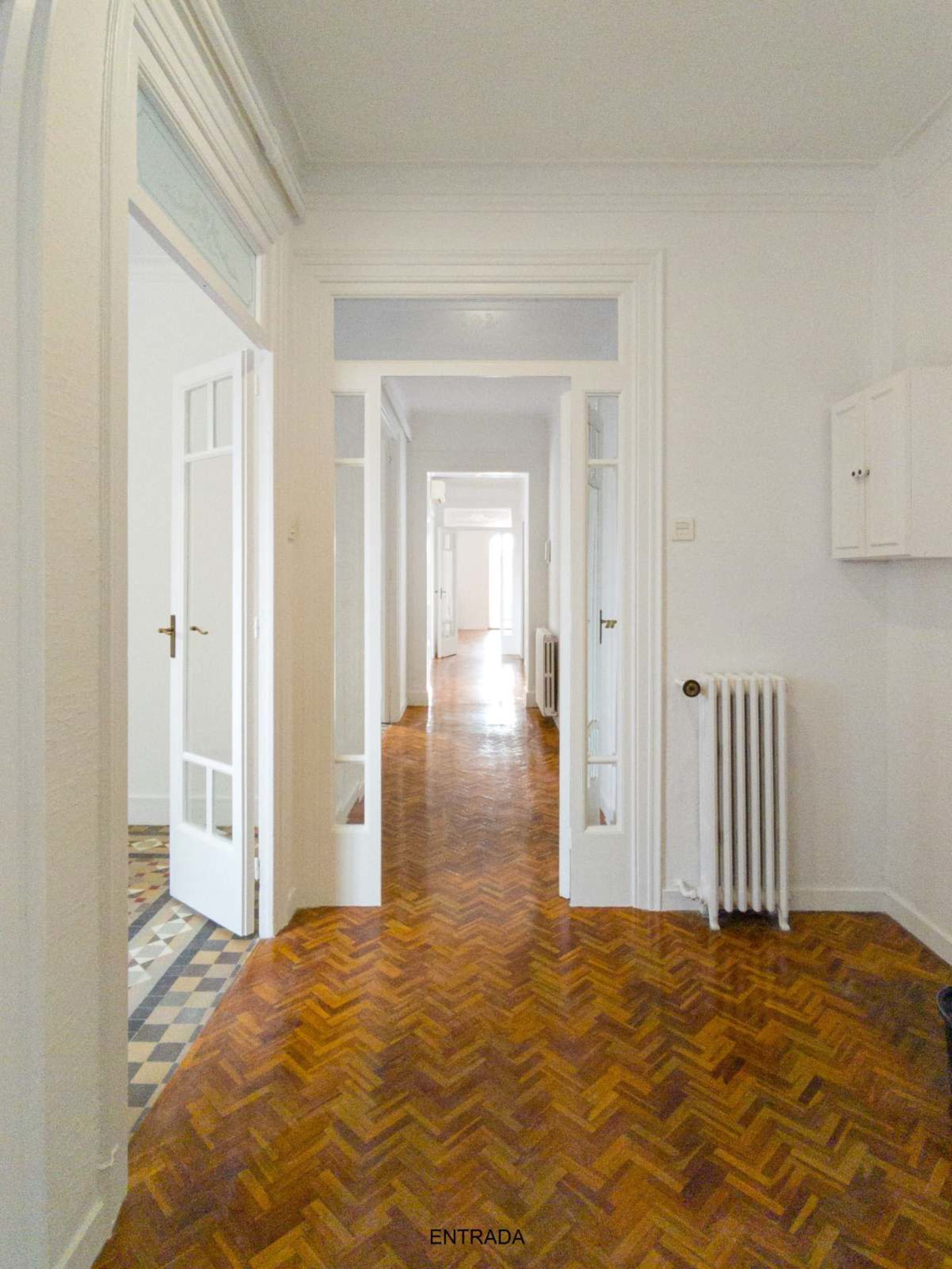 The image shows a long hallway in a well-lit apartment with wooden herringbone flooring and several doorways leading to other rooms.