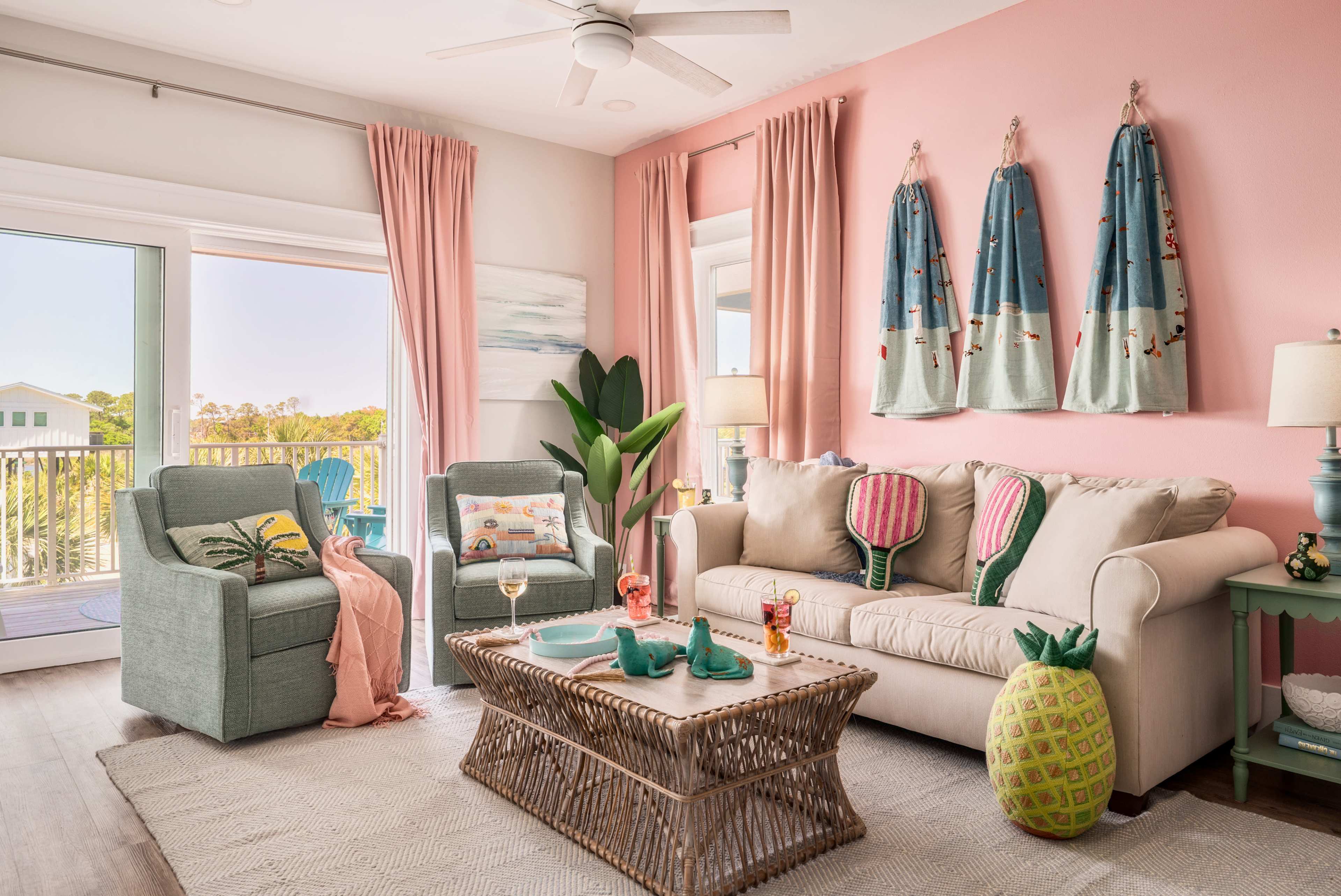 The living room features a light-colored sofa, two chairs, a wicker coffee table, and decorative artwork on the walls, with large windows offering a view of a coastal landscape.