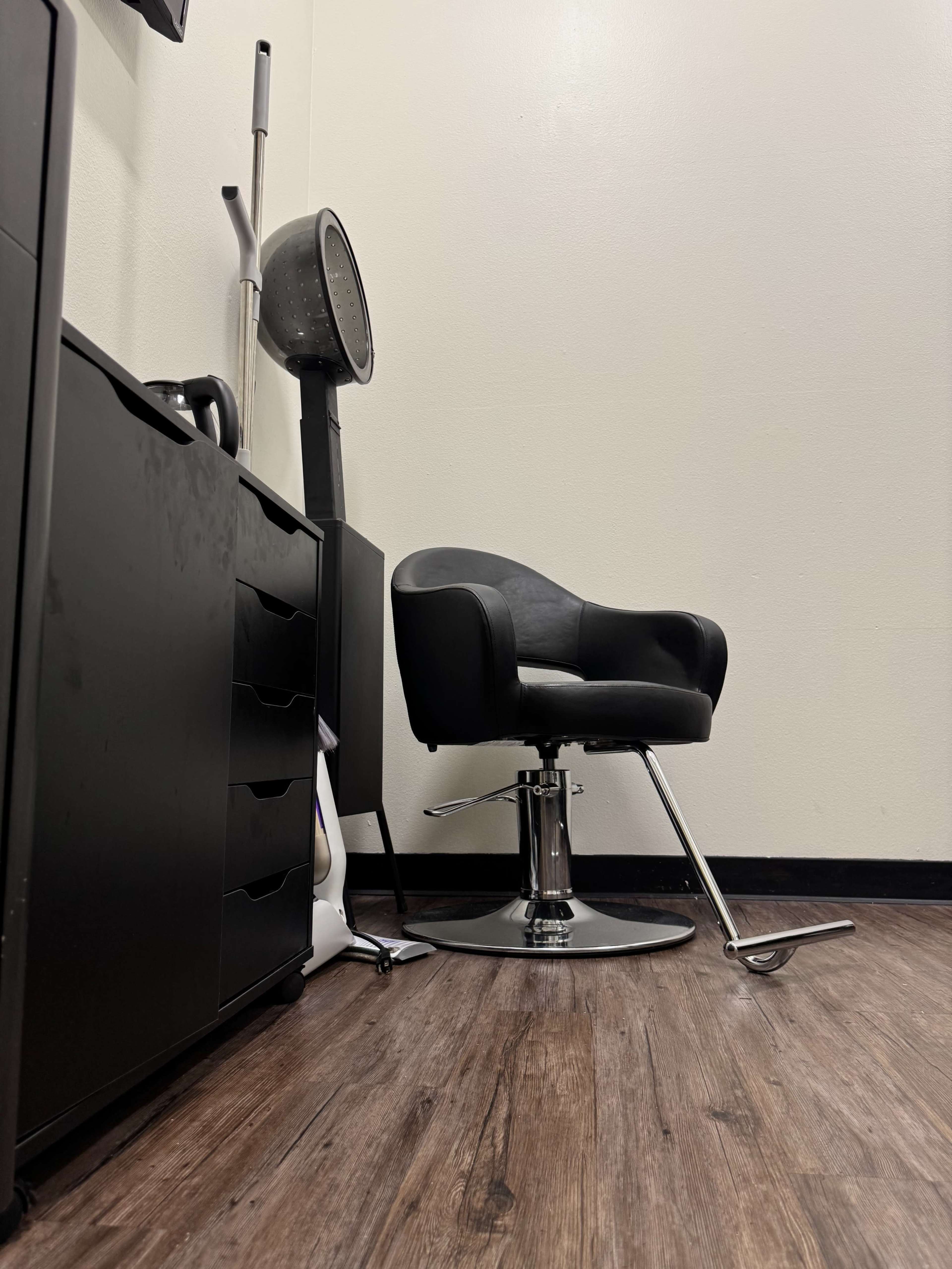 The image shows a modern salon chair in a minimalist setting with a hairdryer on a stand and a cabinet.