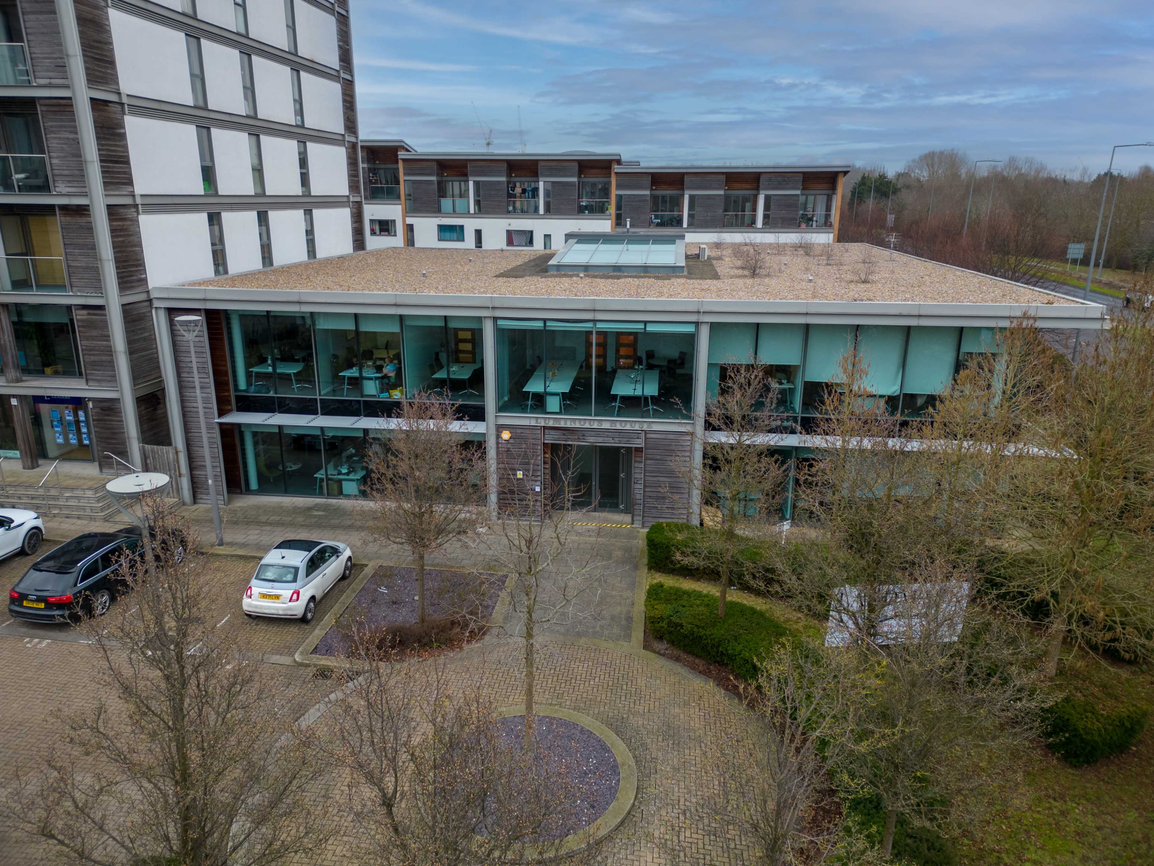 A modern building with large glass windows and a flat roof, surrounded by parked cars and sparse trees.