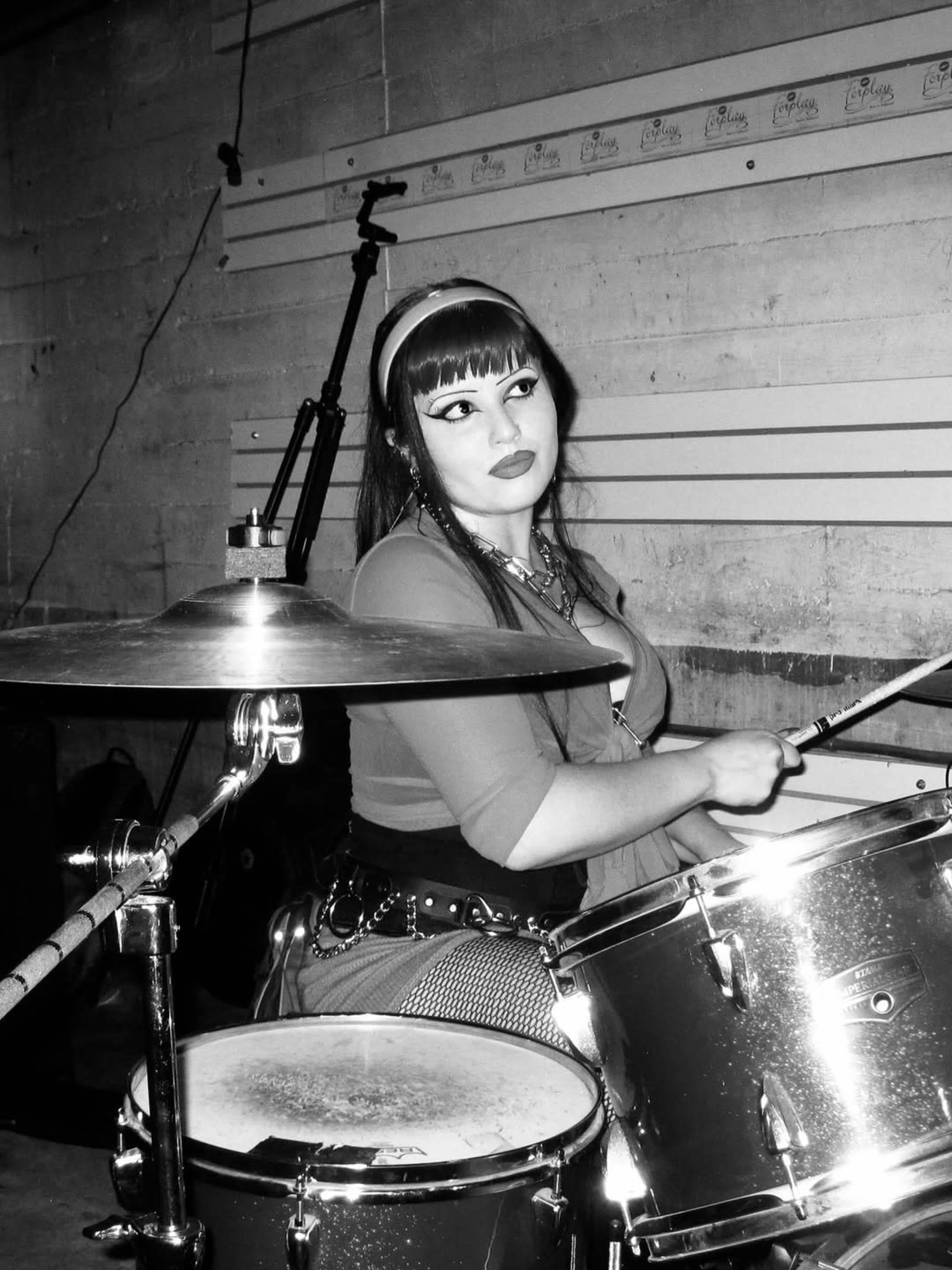 A woman plays the drums in a dimly lit room with exposed concrete walls.