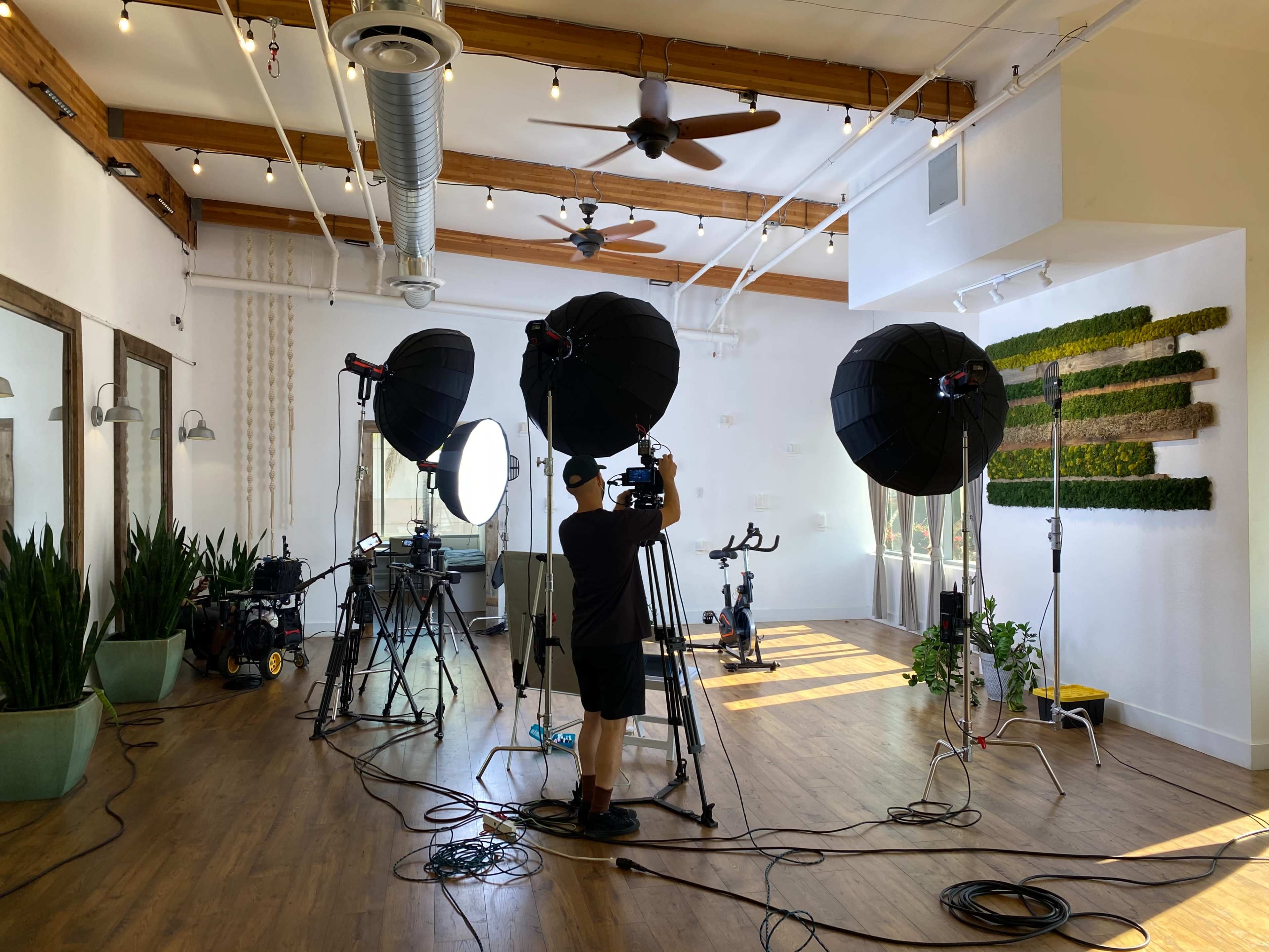 A photographer sets up lighting equipment and cameras in a spacious studio with large windows and greenery on the walls.