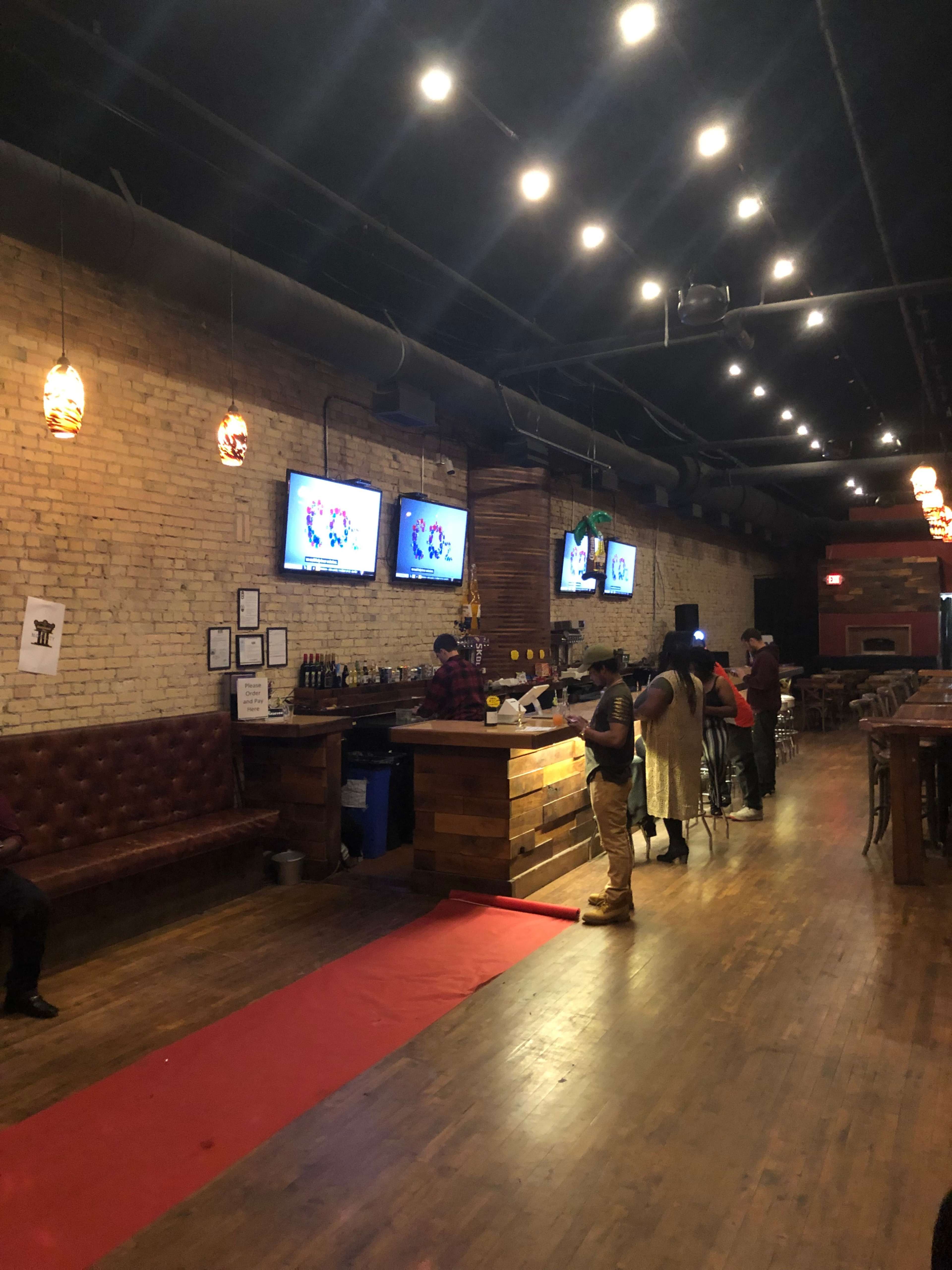 A bar area features a wooden counter, two televisions displaying a program, and patrons interacting on a red carpeted floor under warm lighting.