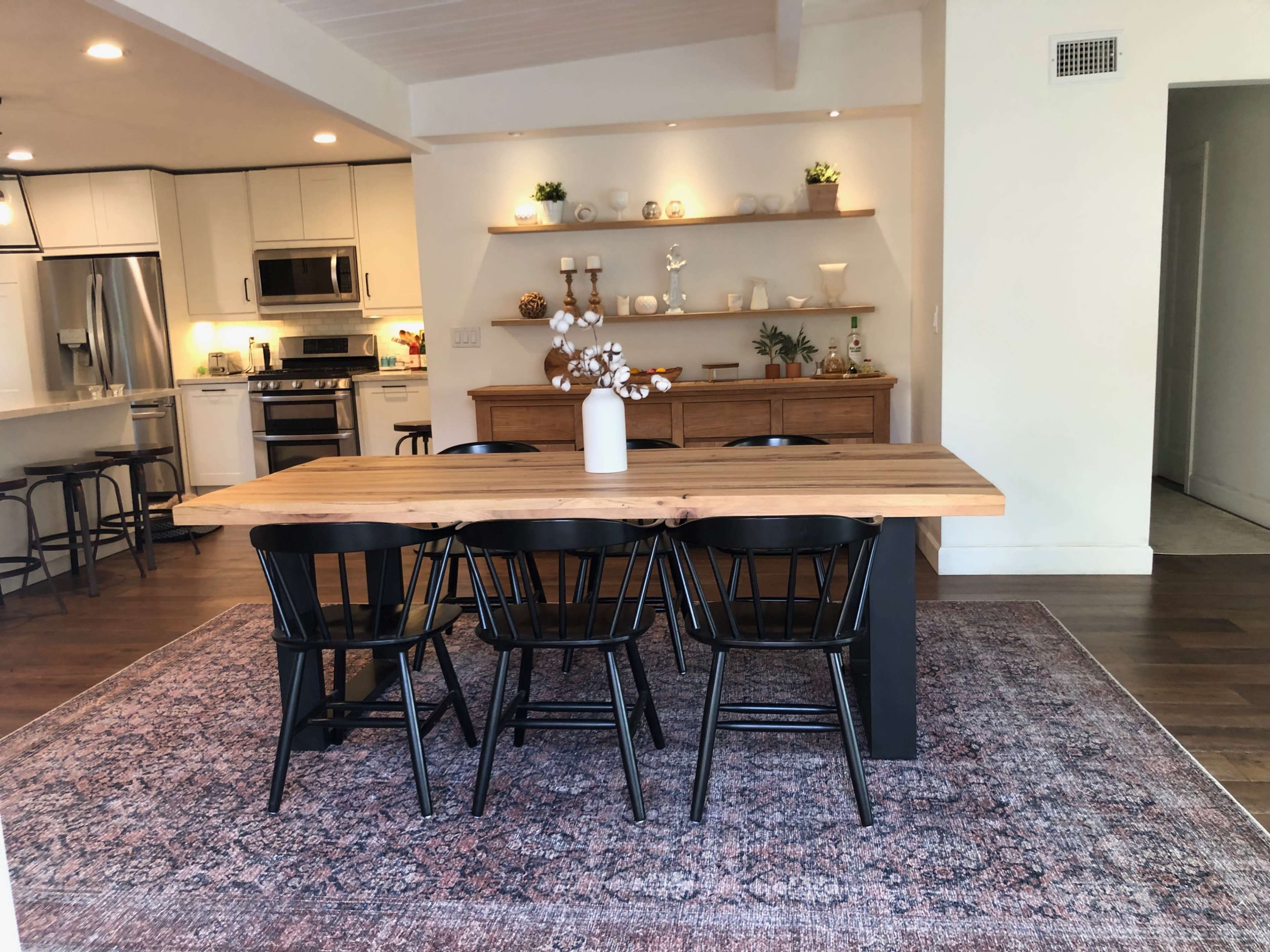 A dining room features a wooden table with four black chairs, surrounded by a patterned rug, and a minimalist wooden shelf displaying decorative items in the background.