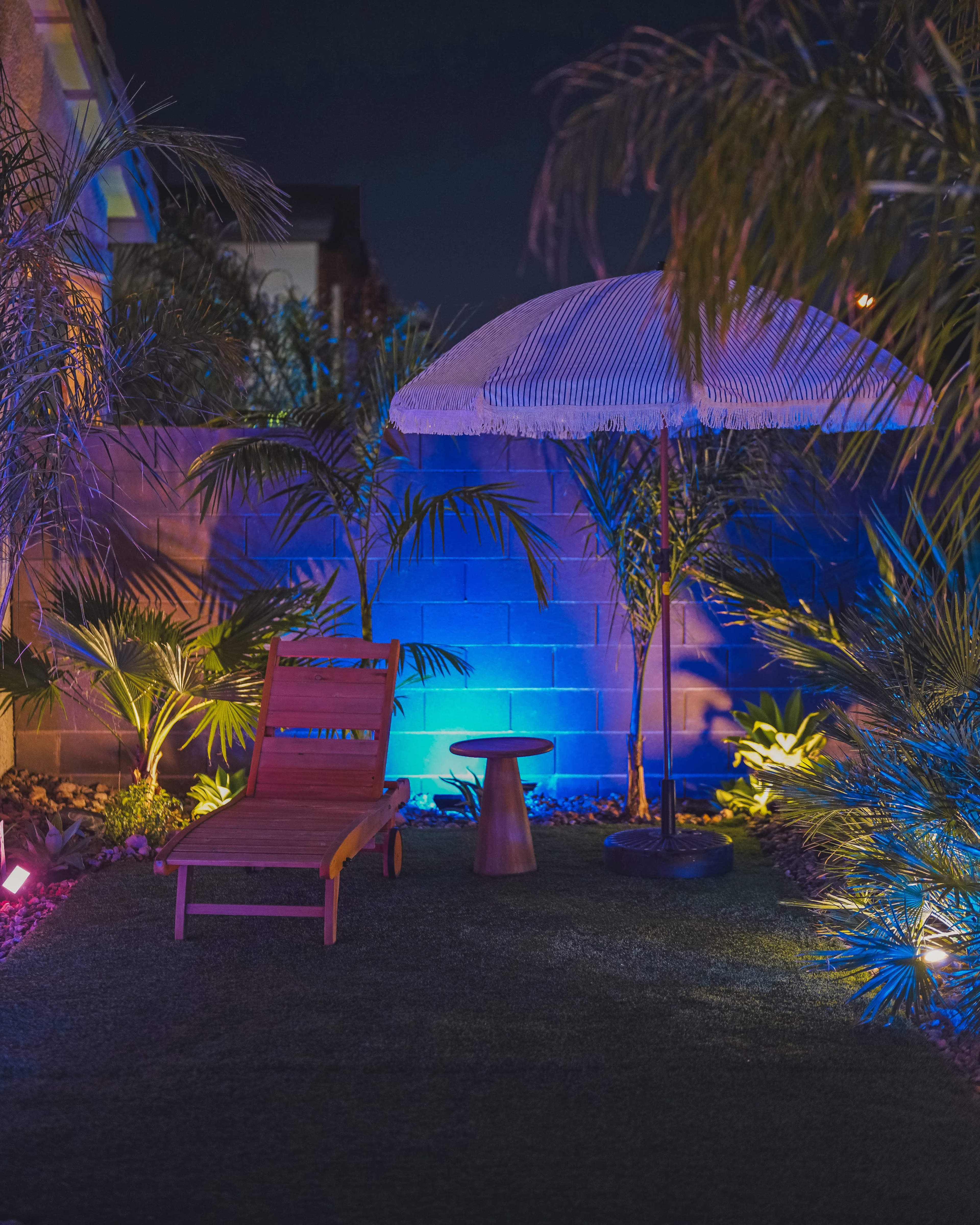 A wooden lounge chair and a small table sit beneath a large umbrella in a landscaped backyard illuminated by blue and purple lights.